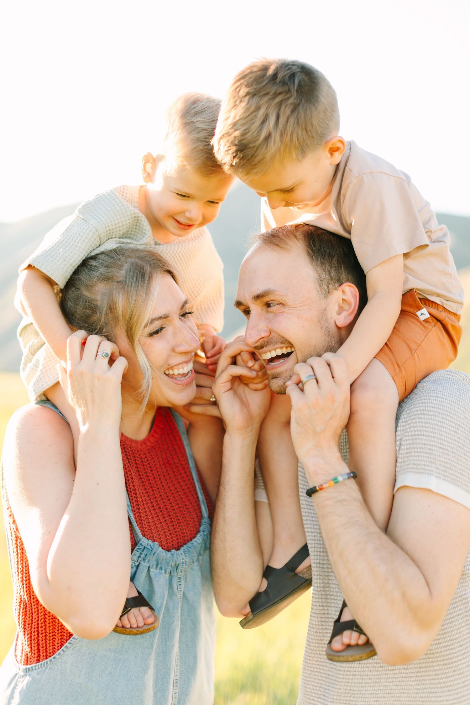 A joyful family with two young boys on their parents shoulders laughing together in a sunny outdoor field.