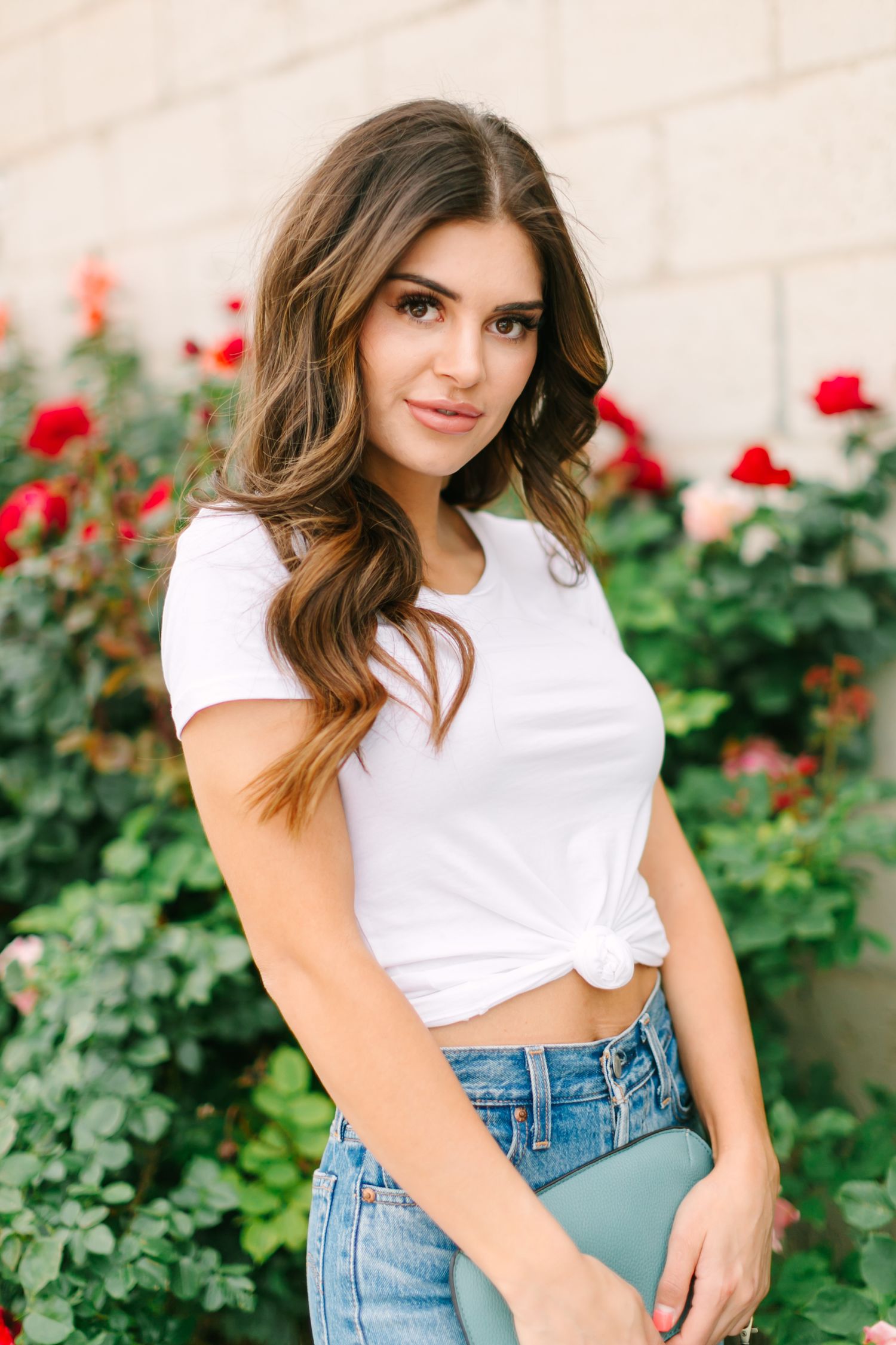 A person in a white crop top and high-waisted jeans poses casually against a wall with red roses in the background.