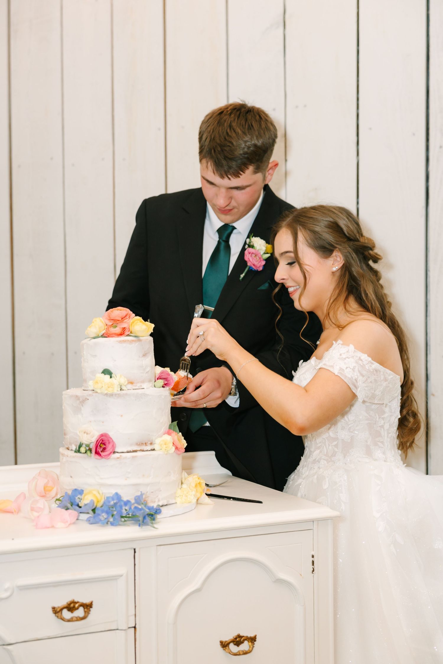 Newlyweds share a playful moment while cutting their three-tiered wedding cake decorated with pink and blue flowers.