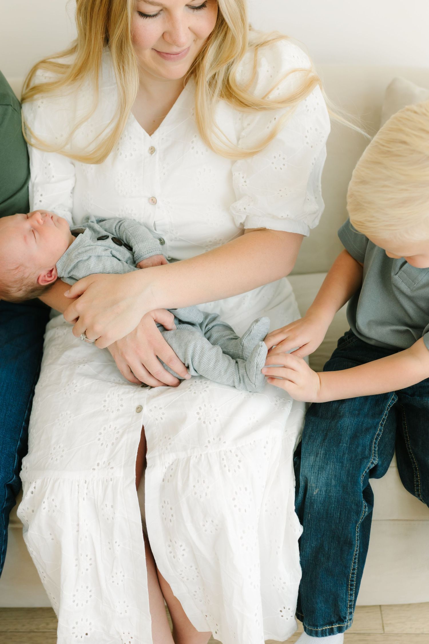 A mother in a white dress holds her newborn baby while an older sibling reaches out to touch the infant.