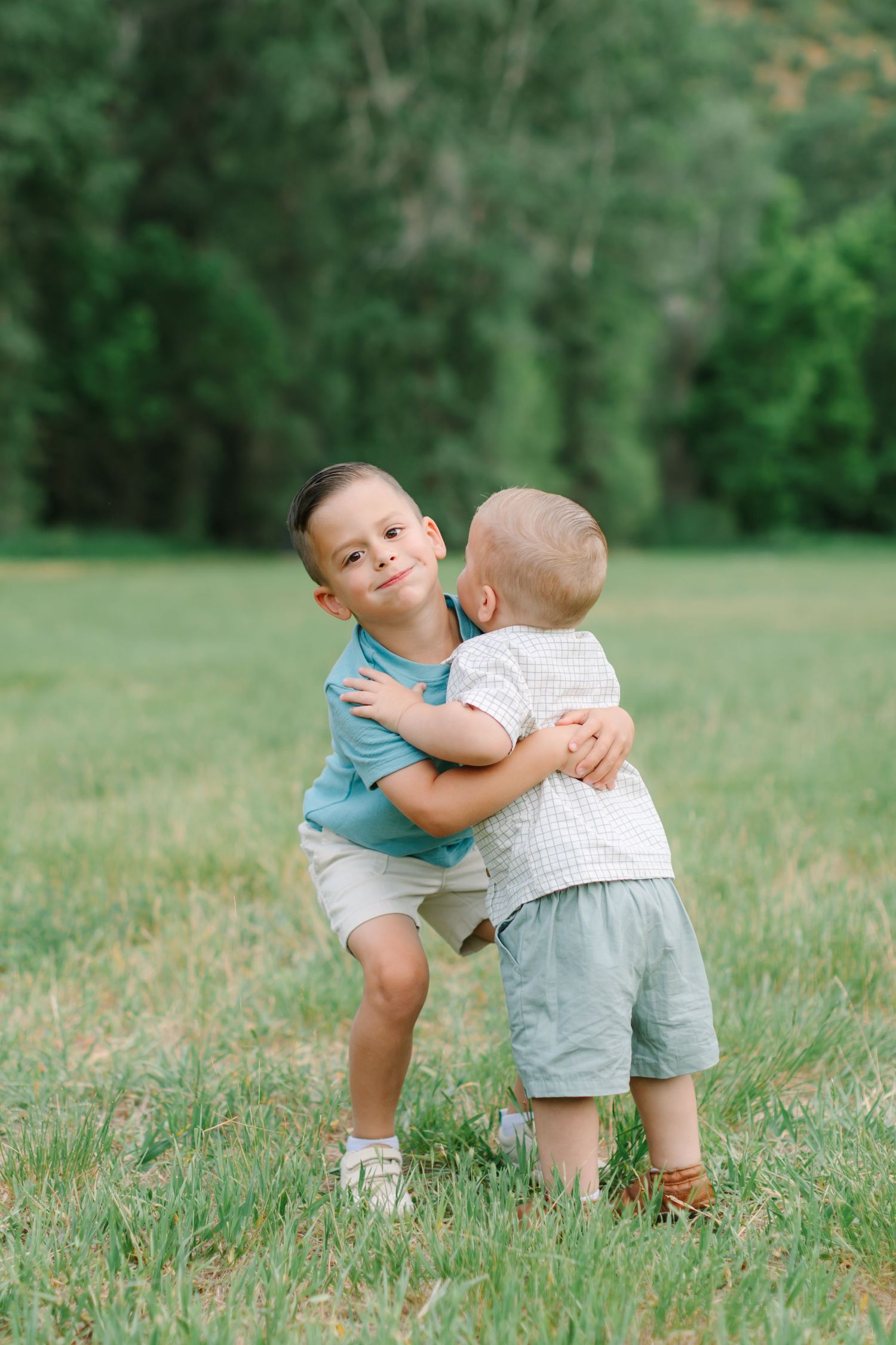Two young children hugging and playing together in a grassy field with green trees in the background.