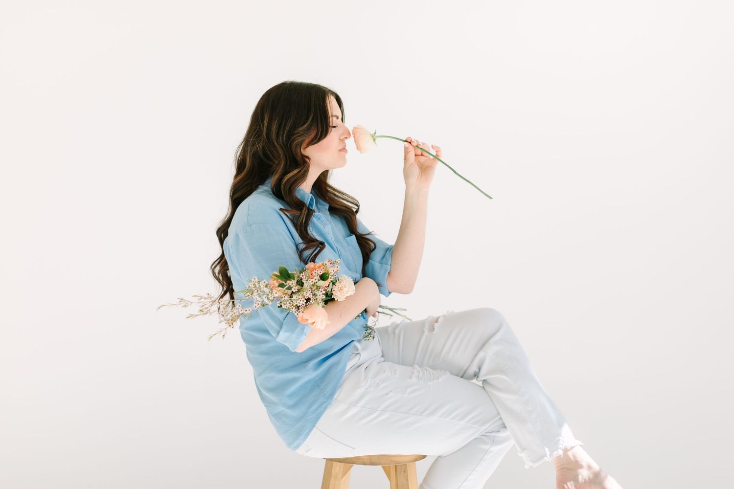 Woman in blue top and white pants sits casually in studio setting while writing in notebook.