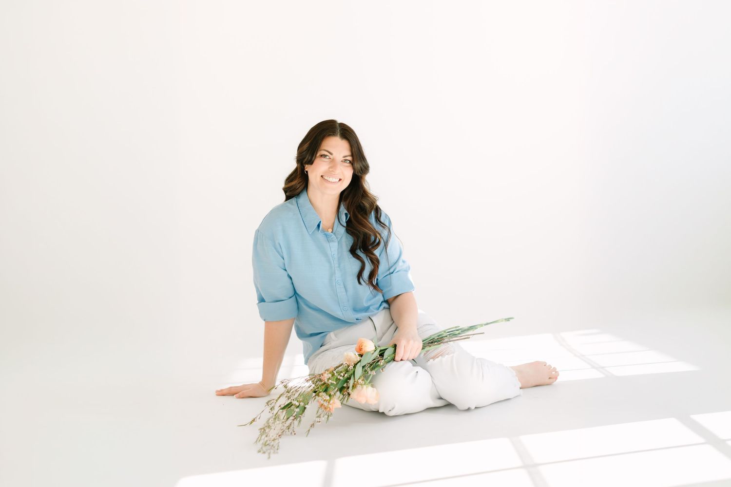 Person in light blue shirt lies on white floor with floral arrangement and ceramic bowl in minimalist setting.