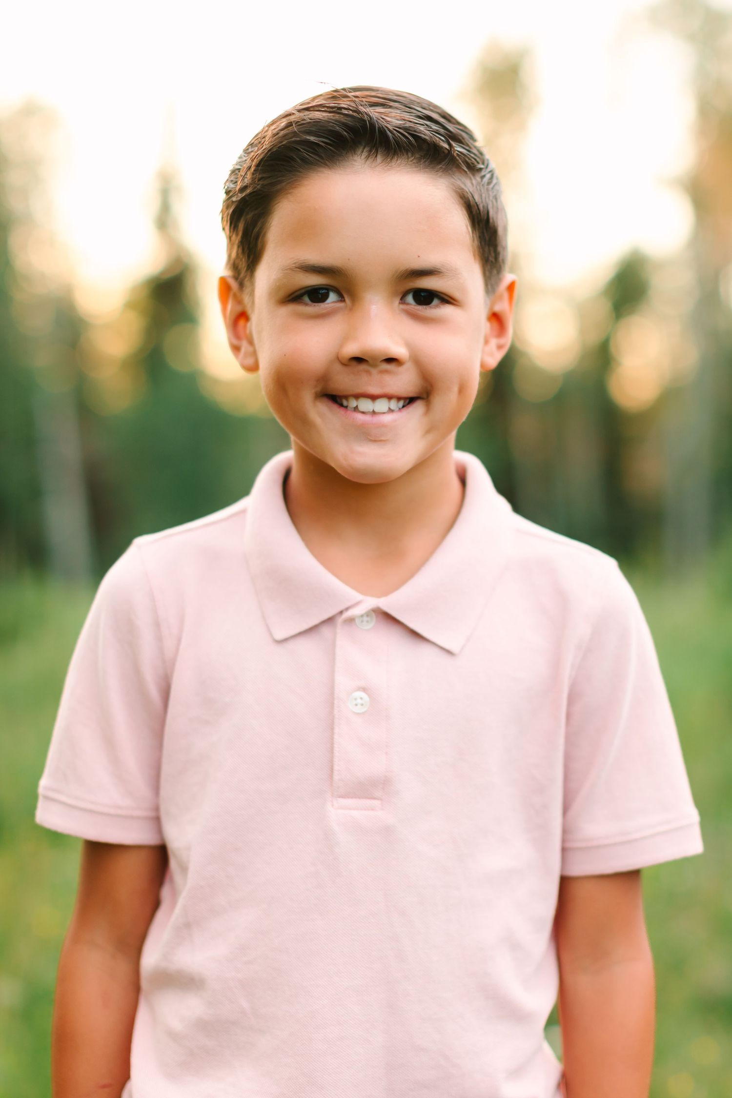 Smiling young boy in a pink polo shirt posing outdoors with green trees in the background.