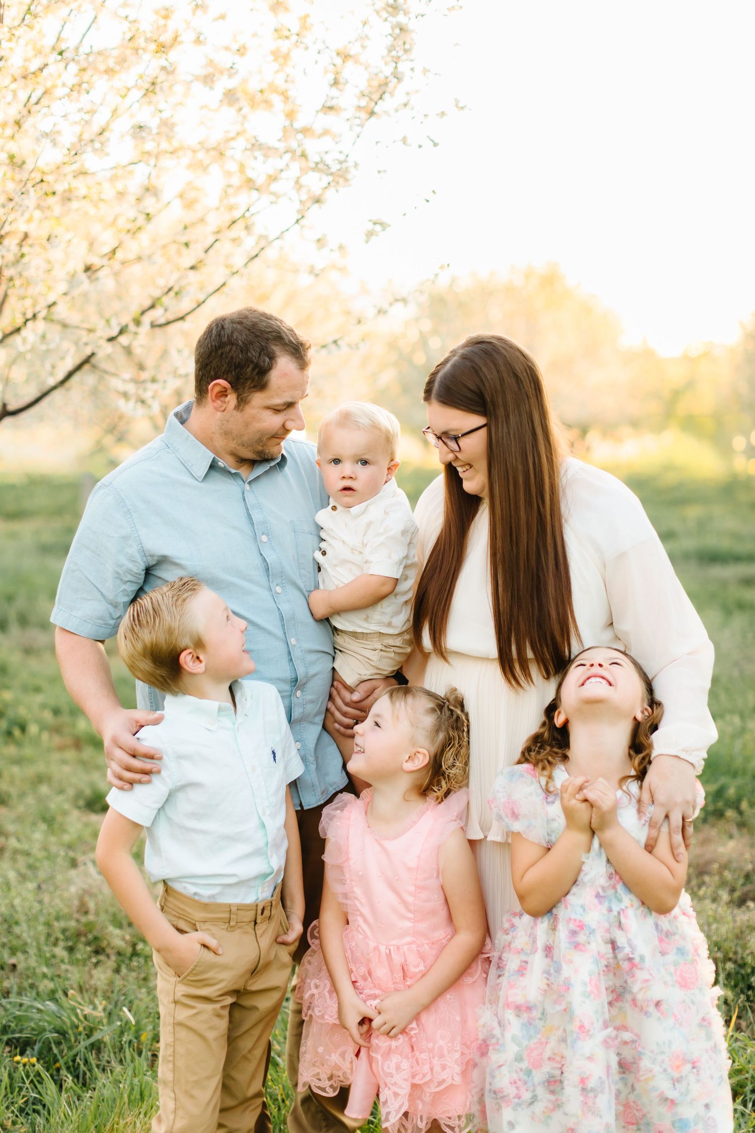Family of six posing outdoors near blooming trees in golden light, parents smiling at baby while children laugh and play.