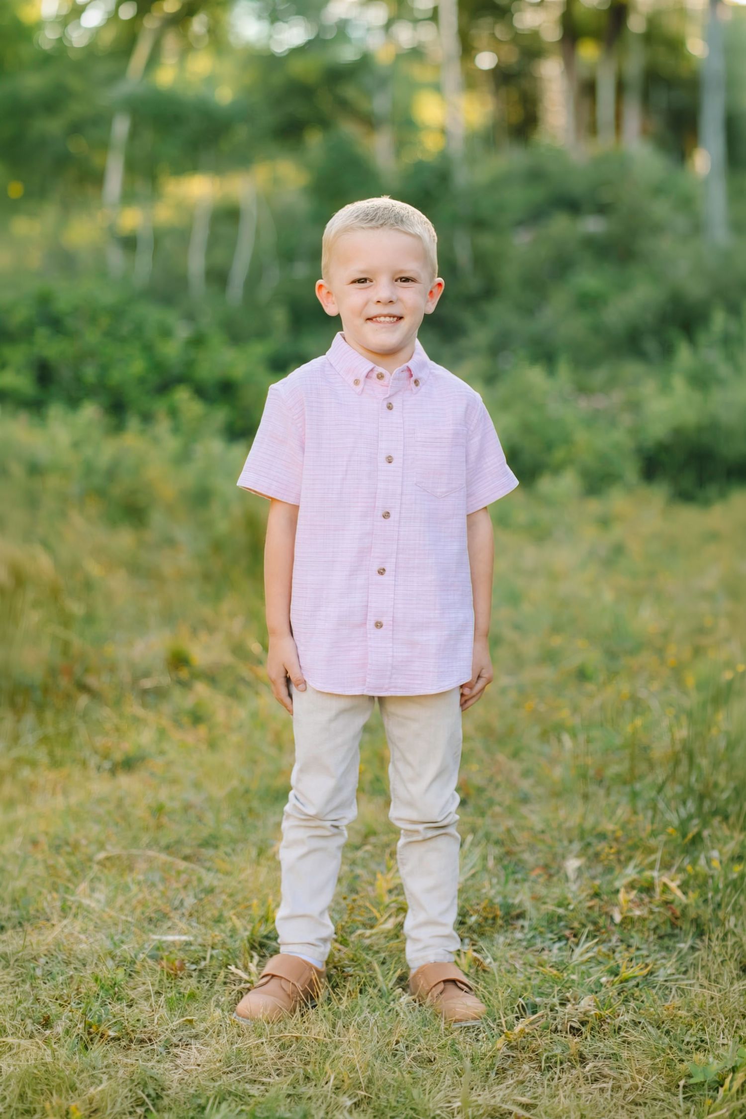 A smiling young blonde boy in a pink button-up shirt and khaki pants stands outdoors in a green field in American Fork, Utah.