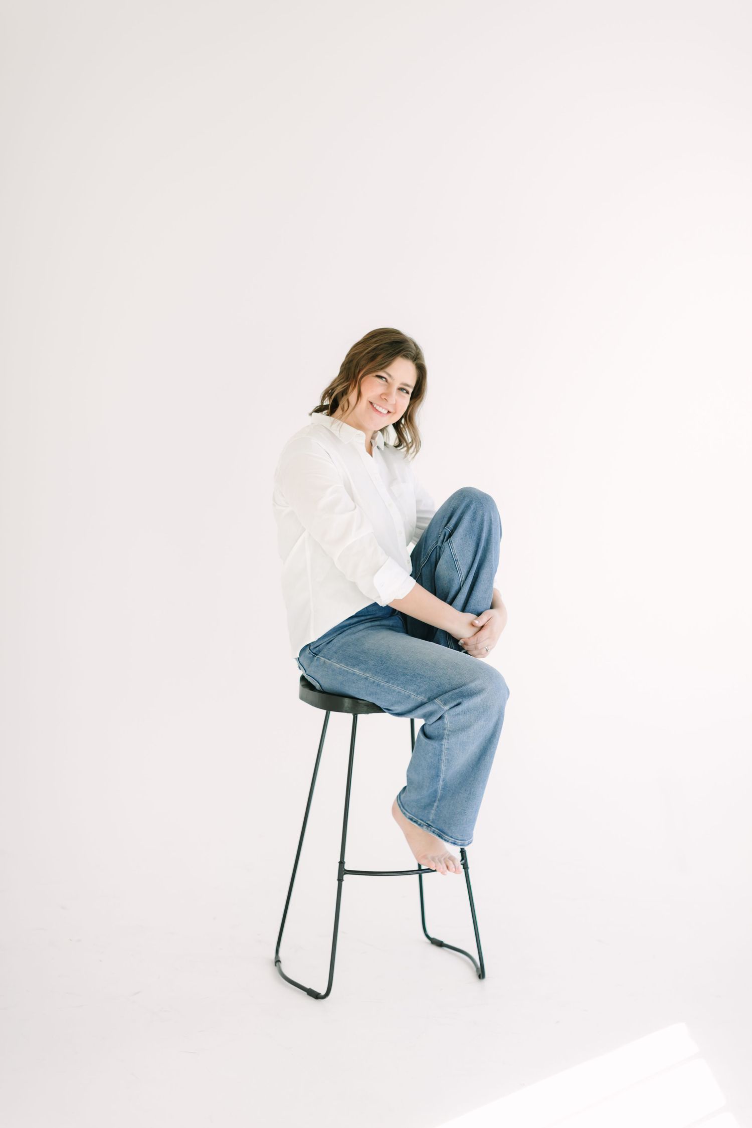 Person in casual white shirt and jeans sits perched on a minimalist black metal barstool against white background.