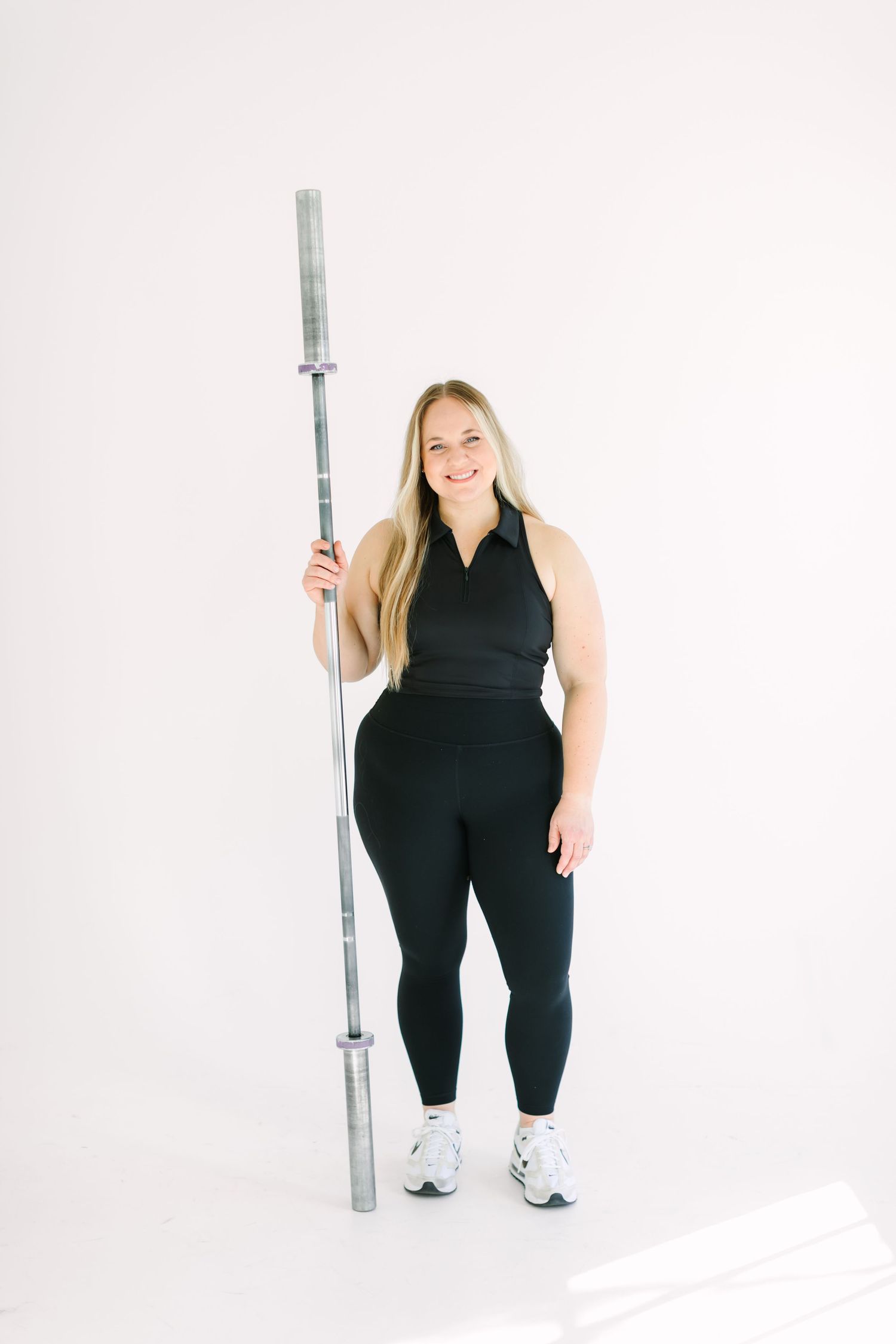 Person in black athletic wear stands confidently against white background holding a silver pole.