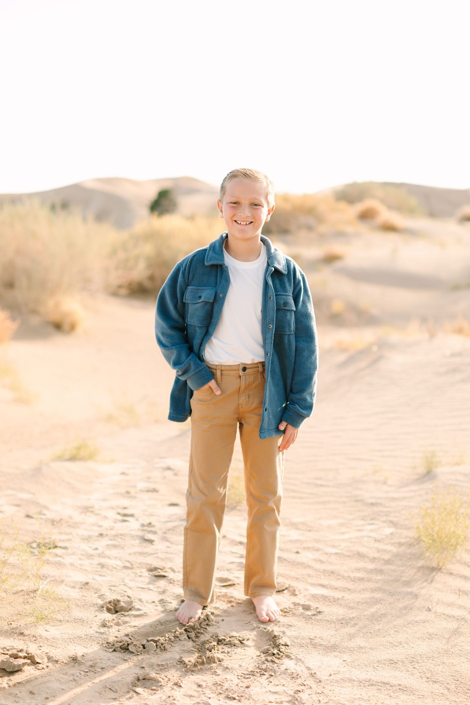Small child wearing denim jacket and khaki pants plays on sandy desert terrain at sunset.