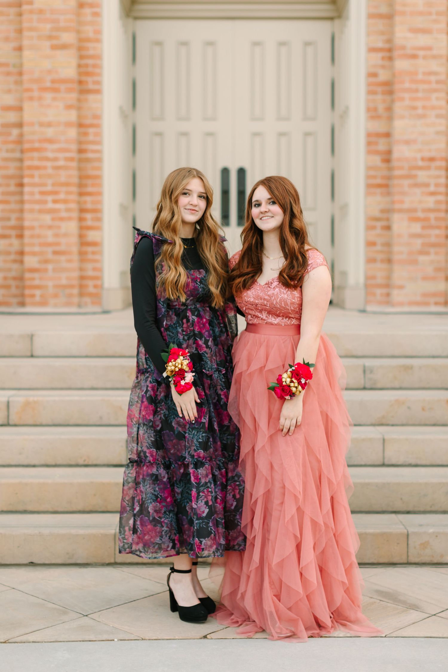 Two people pose together in complementary dresses, one in dark floral and one in coral pink, on stone steps.