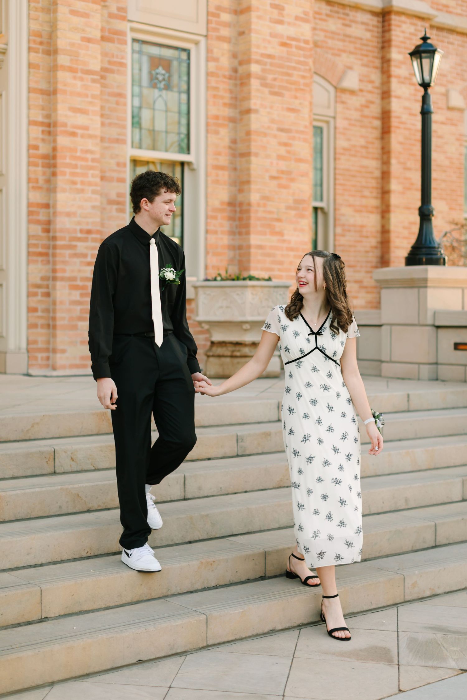 A couple in formal attire stand on stone steps outside a brick building with a vintage lamp post.