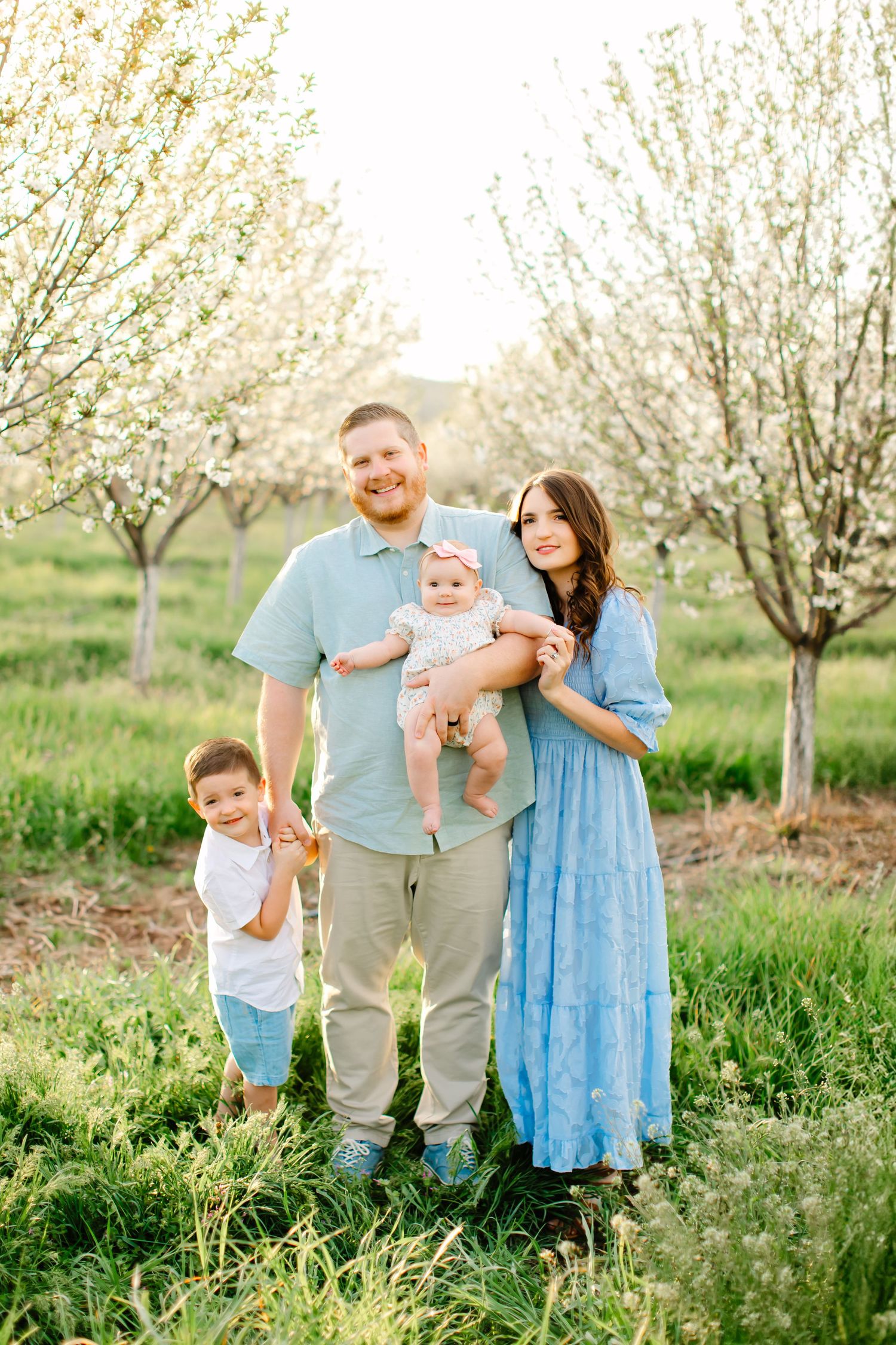 A family plays together in green grass at a blooming orchard for their family photoshoot in Santaquin, Utah.