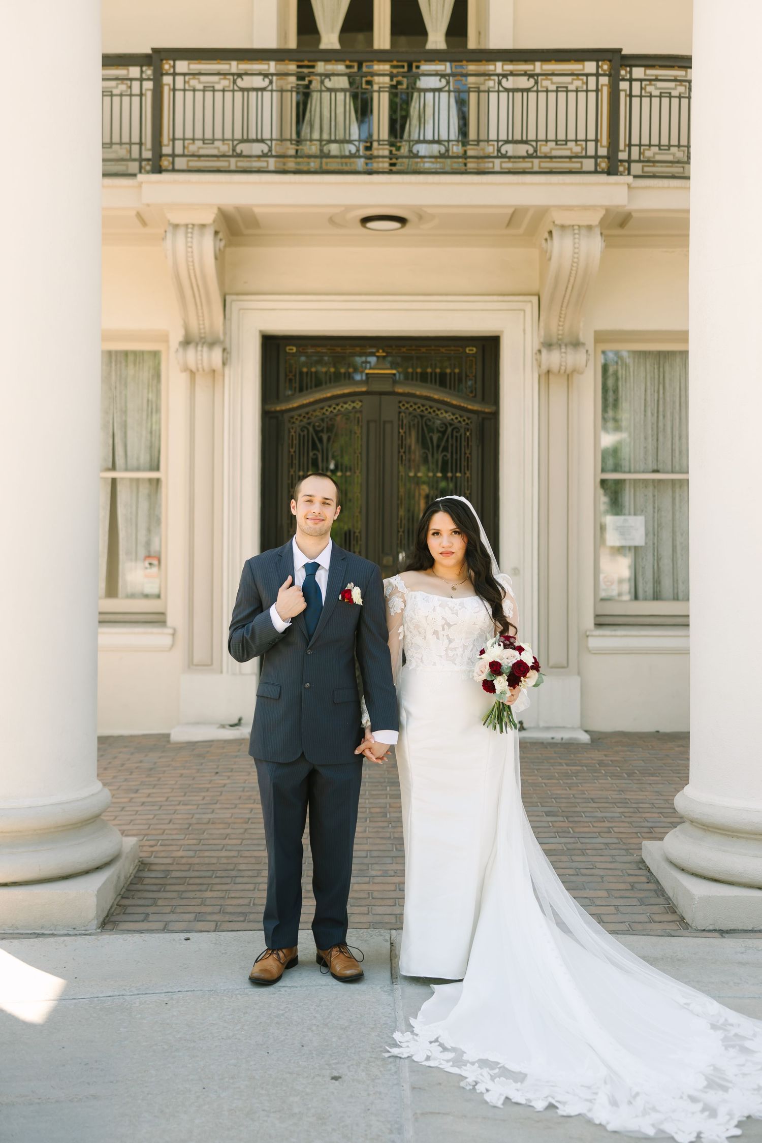 A bride and groom pose together in front of an elegant white columned entrance with grand architecture.