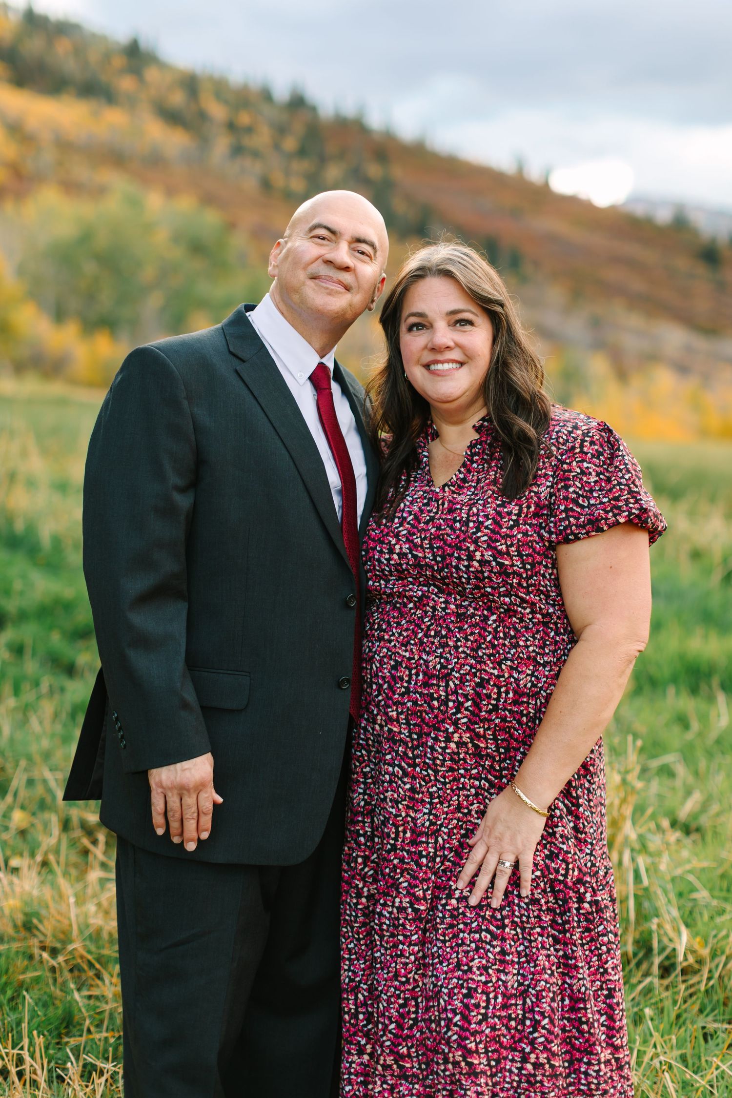 A couple poses together in an autumn field with golden foliage in the background wearing formal attire.