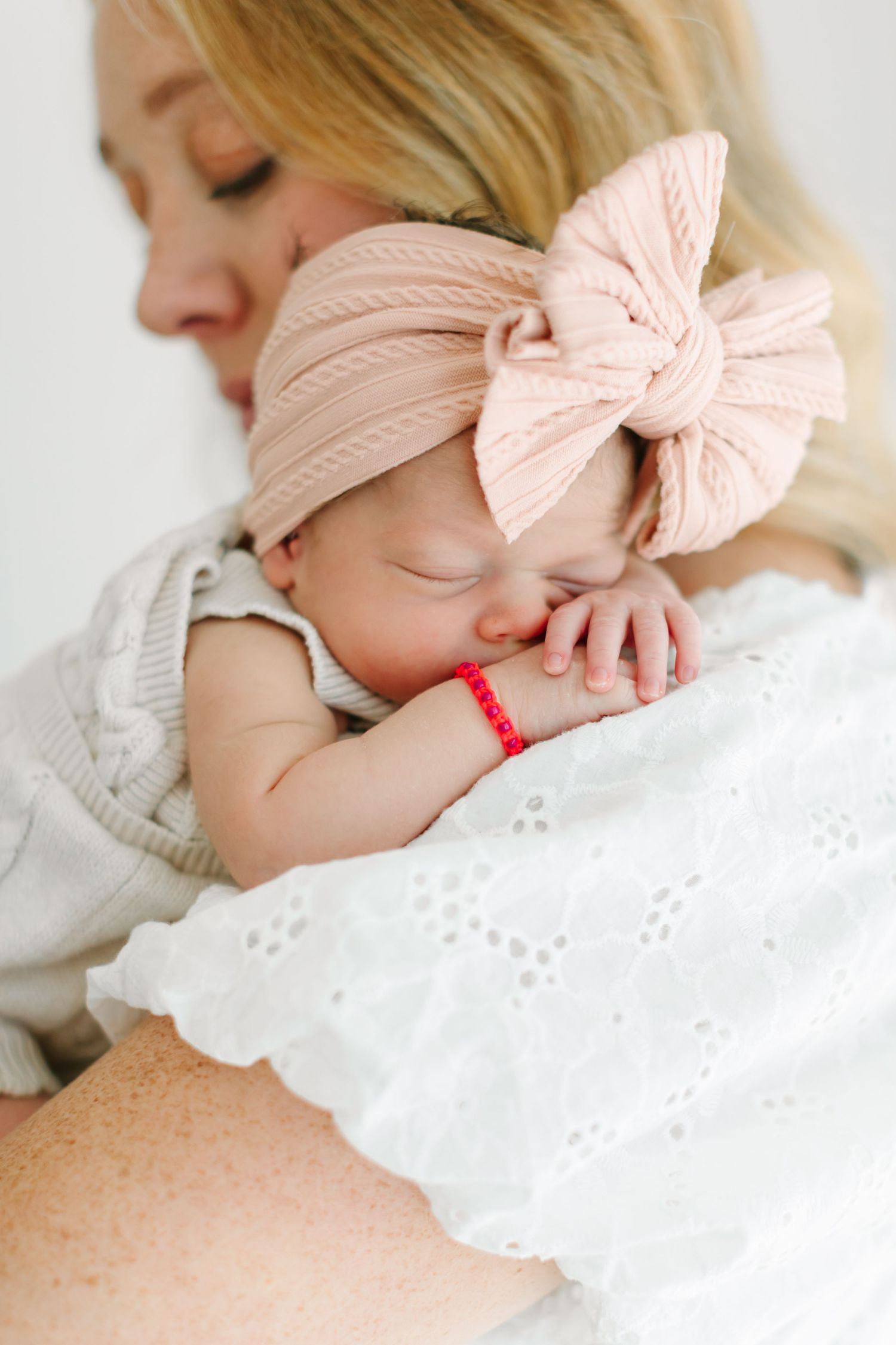 A sleeping newborn wearing a pink bow headband rests peacefully against someone's shoulder.