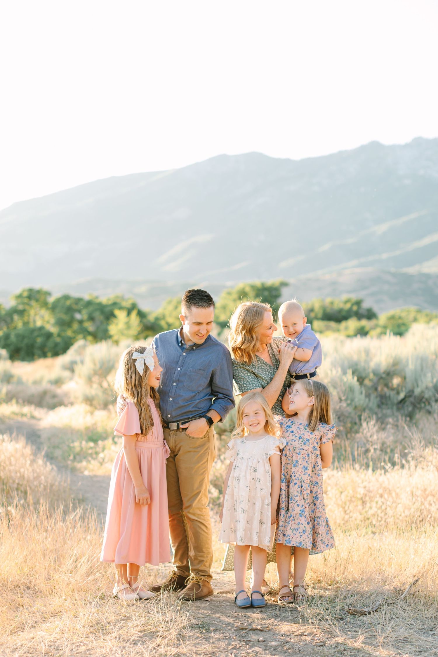 A family shares tender moments together in a golden field with mountains in the background at sunset.