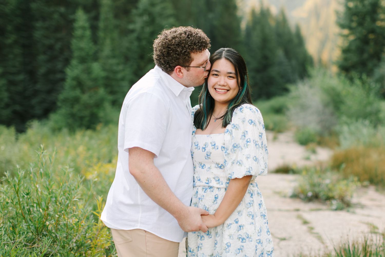 Couple embracing outdoors in a mountain meadow, man kissing woman's cheek as she smiles, surrounded by evergreen trees in Utah.