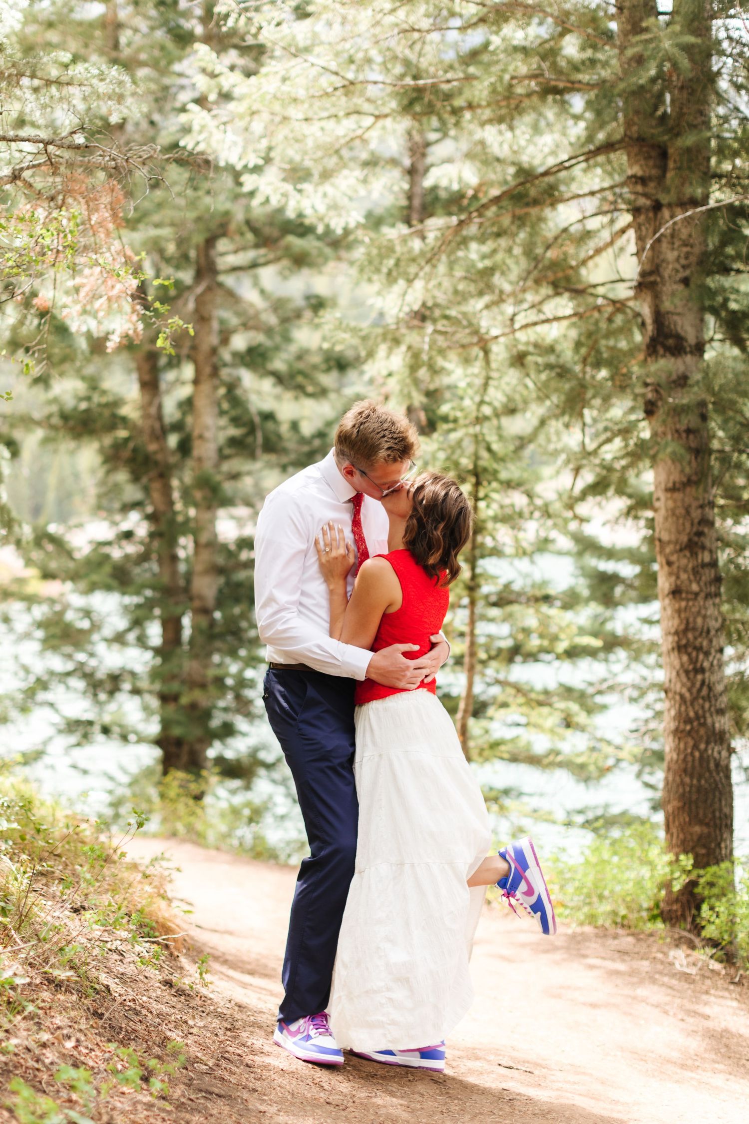 Couple sharing a romantic kiss on a forest trail, bride in red top and white skirt with colorful sneakers.