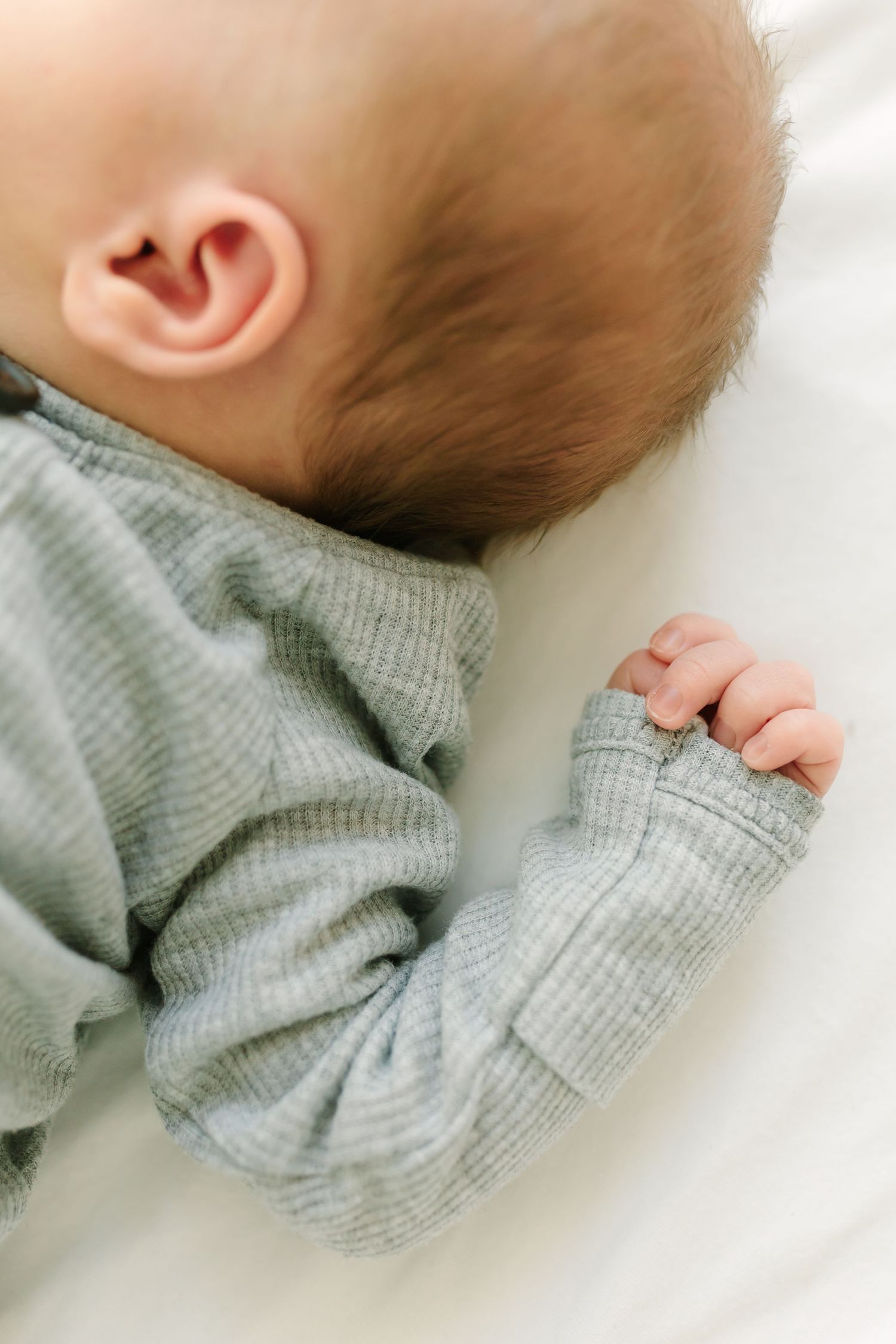 A sleeping baby in a gray ribbed onesie lies on a white surface, showing their tiny hand and ear.