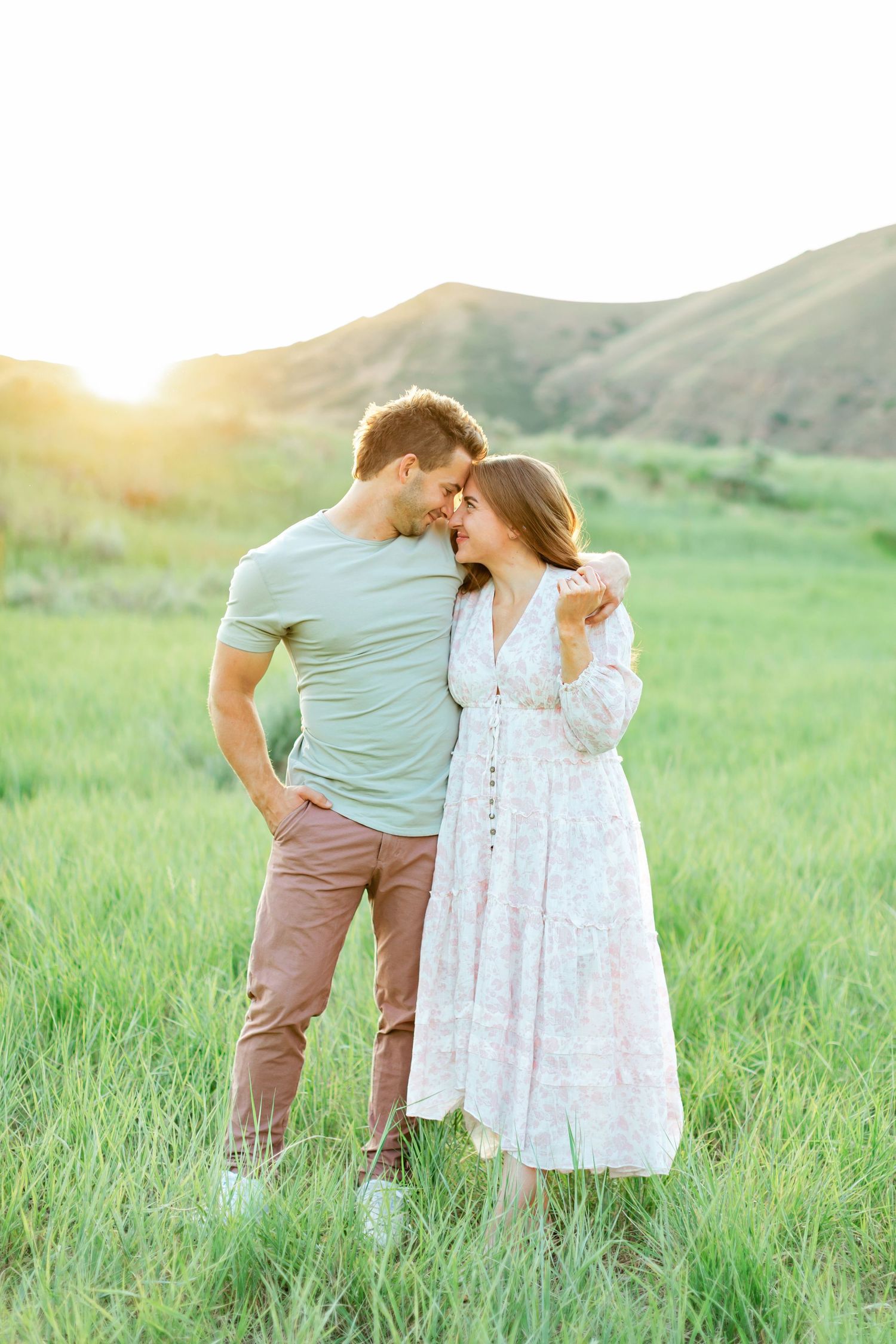 A romantic couple in casual attire shares tender moments in a sunlit grassy meadow with rolling hills in the background.