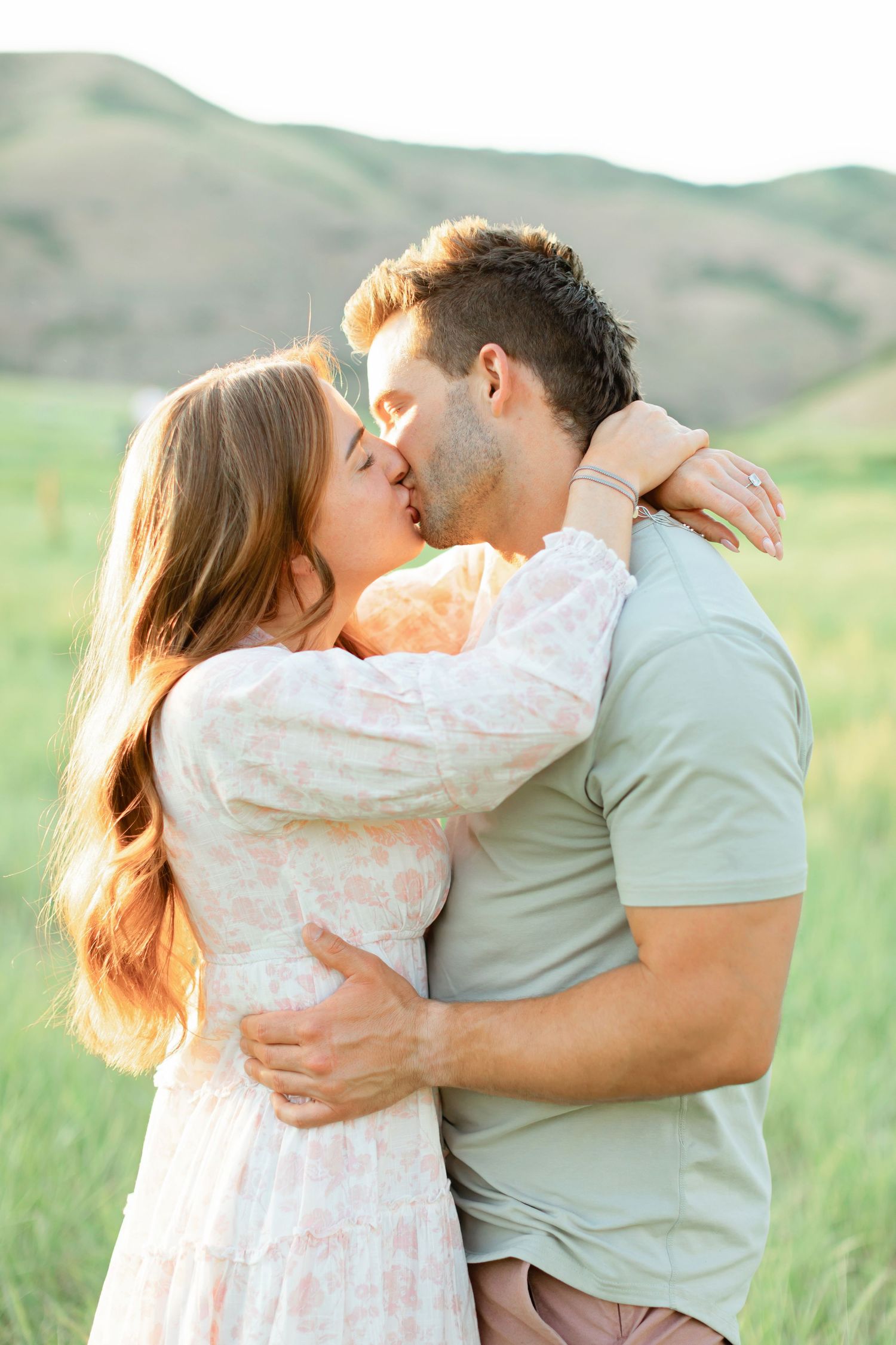 Romantic couple embracing in a scenic mountain meadow wearing soft pastel colors at sunset.