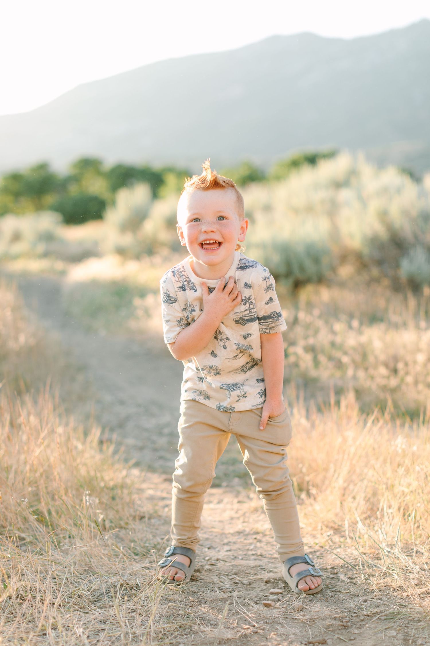 Young child plays and laughs joyfully in a golden field during sunset with mountains in the background.