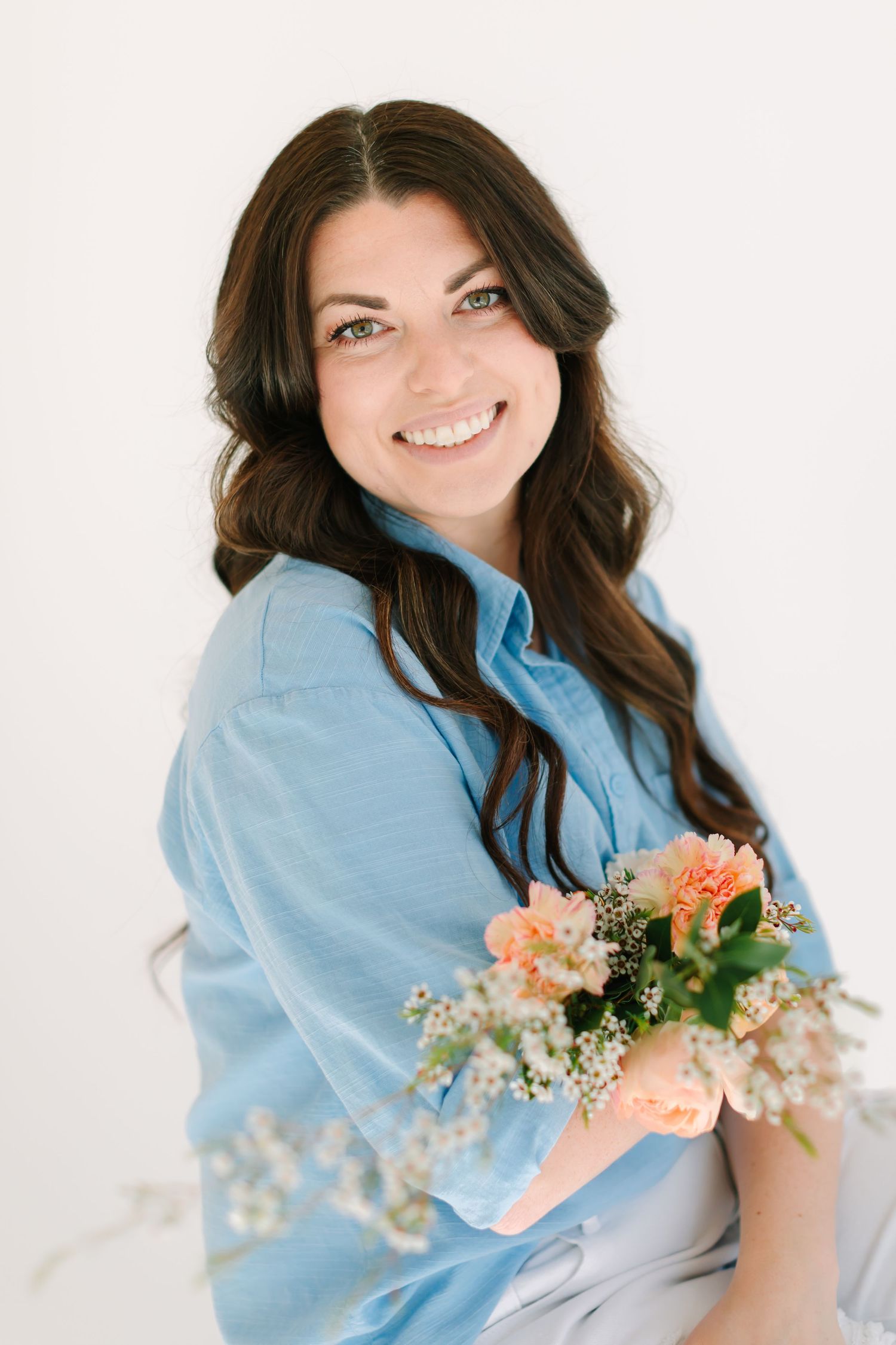 Photographer in light blue shirt holds delicate peach and white flower bouquet against white background.