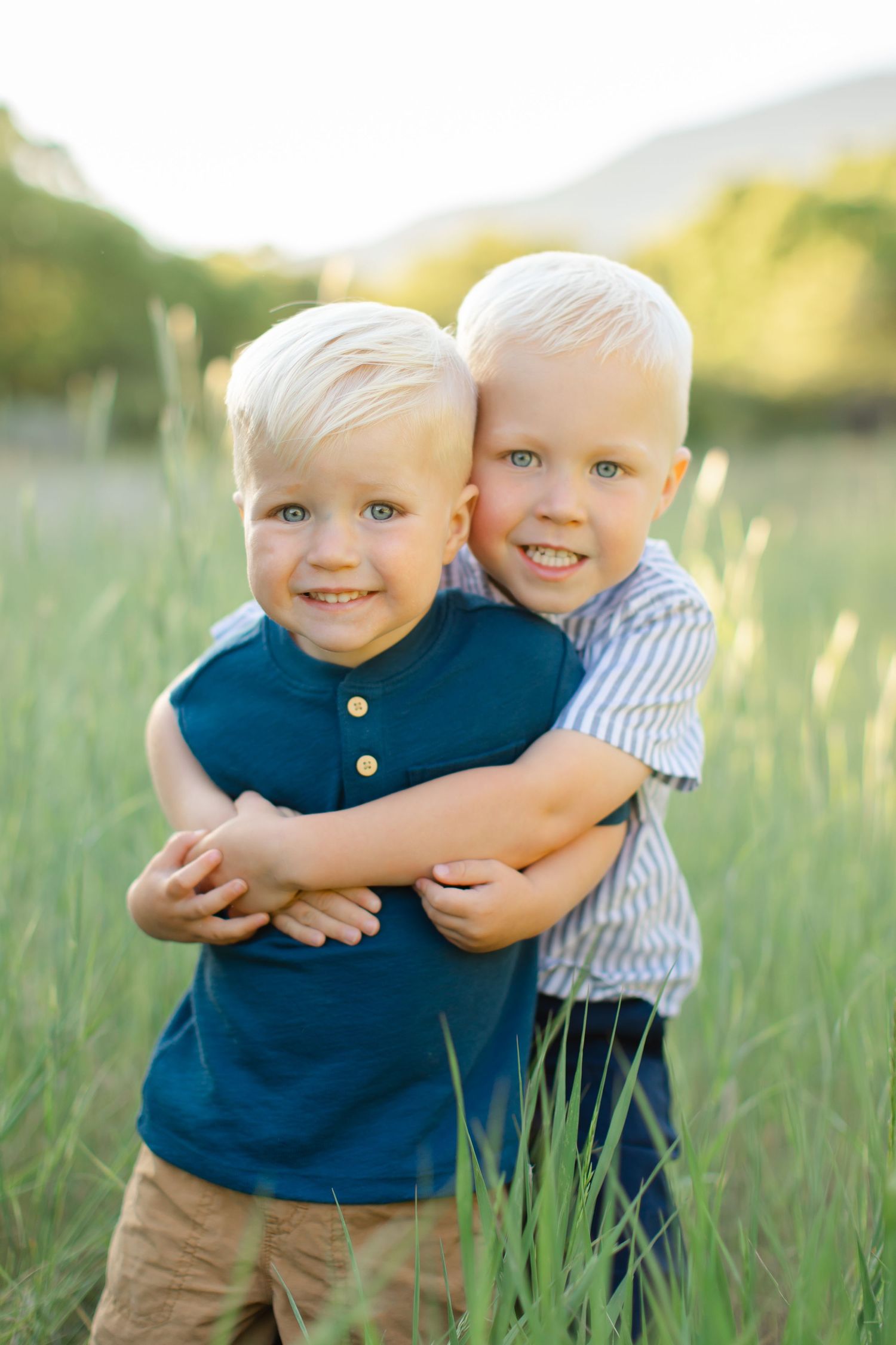 Two young children hugging and smiling together in a grassy field during golden hour.