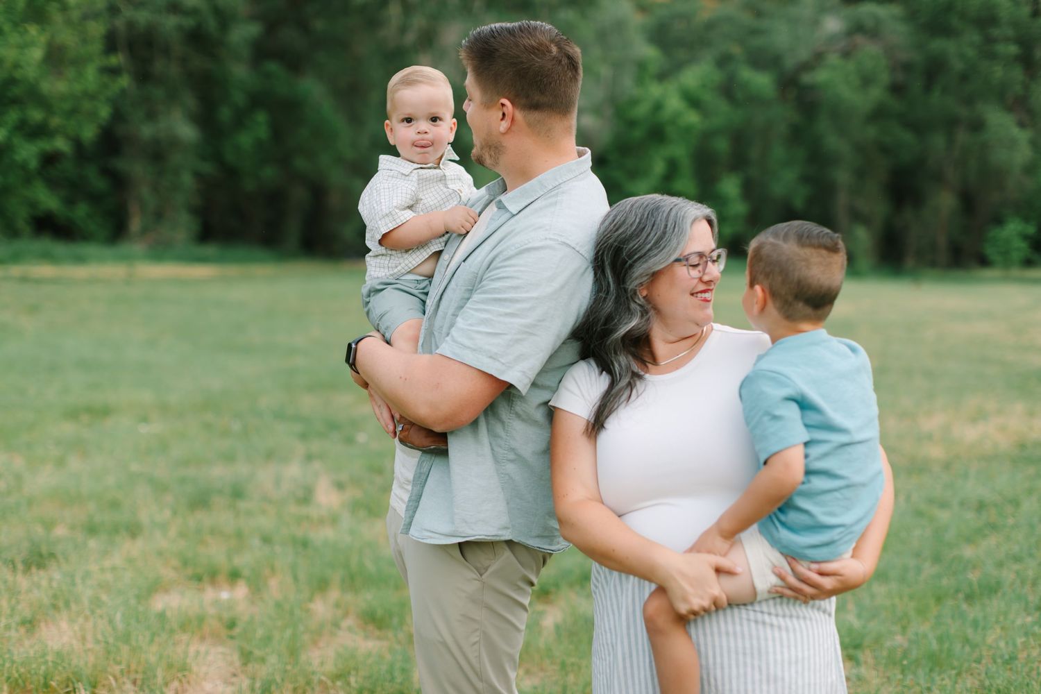 Family wearing coordinated grey and blue outfits embrace together in a sunny green field during outdoor photo session.