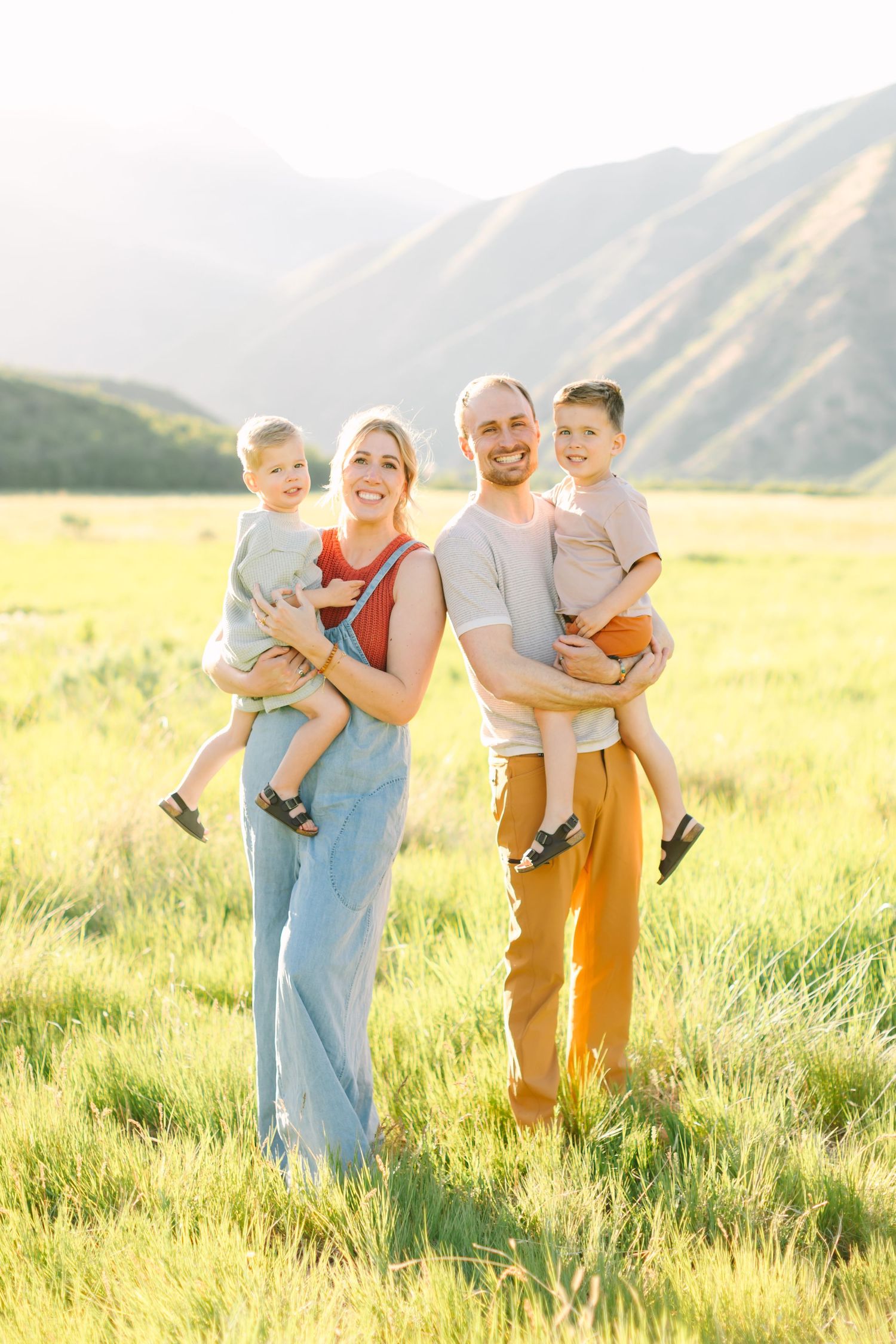 Happy family of four poses in a sunlit green meadow with mountain backdrop, parents holding two young children.