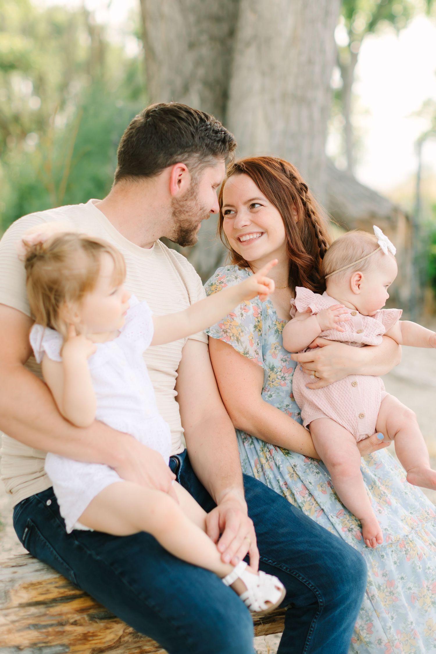 A happy young family with two baby girls sits outdoors, sharing a tender moment together in natural light.