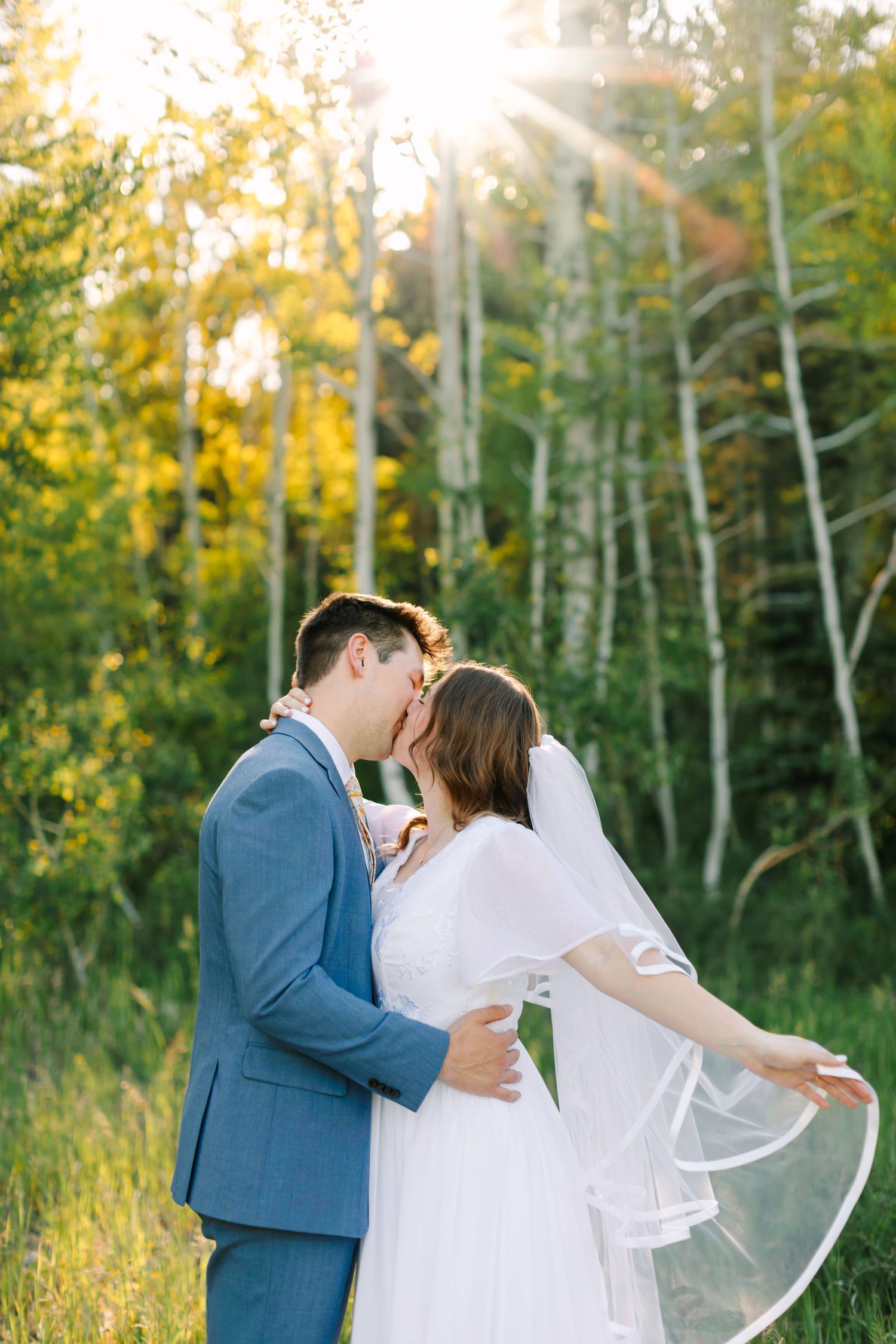 A bride and groom share a romantic kiss outdoors in a sunlit forest, with the veil flowing gracefully in the breeze.
