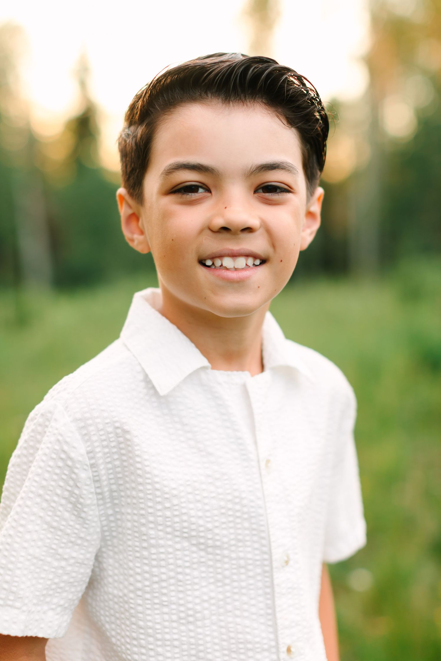 Smiling young boy in a white shirt posing outdoors with green trees in the background during golden hour.