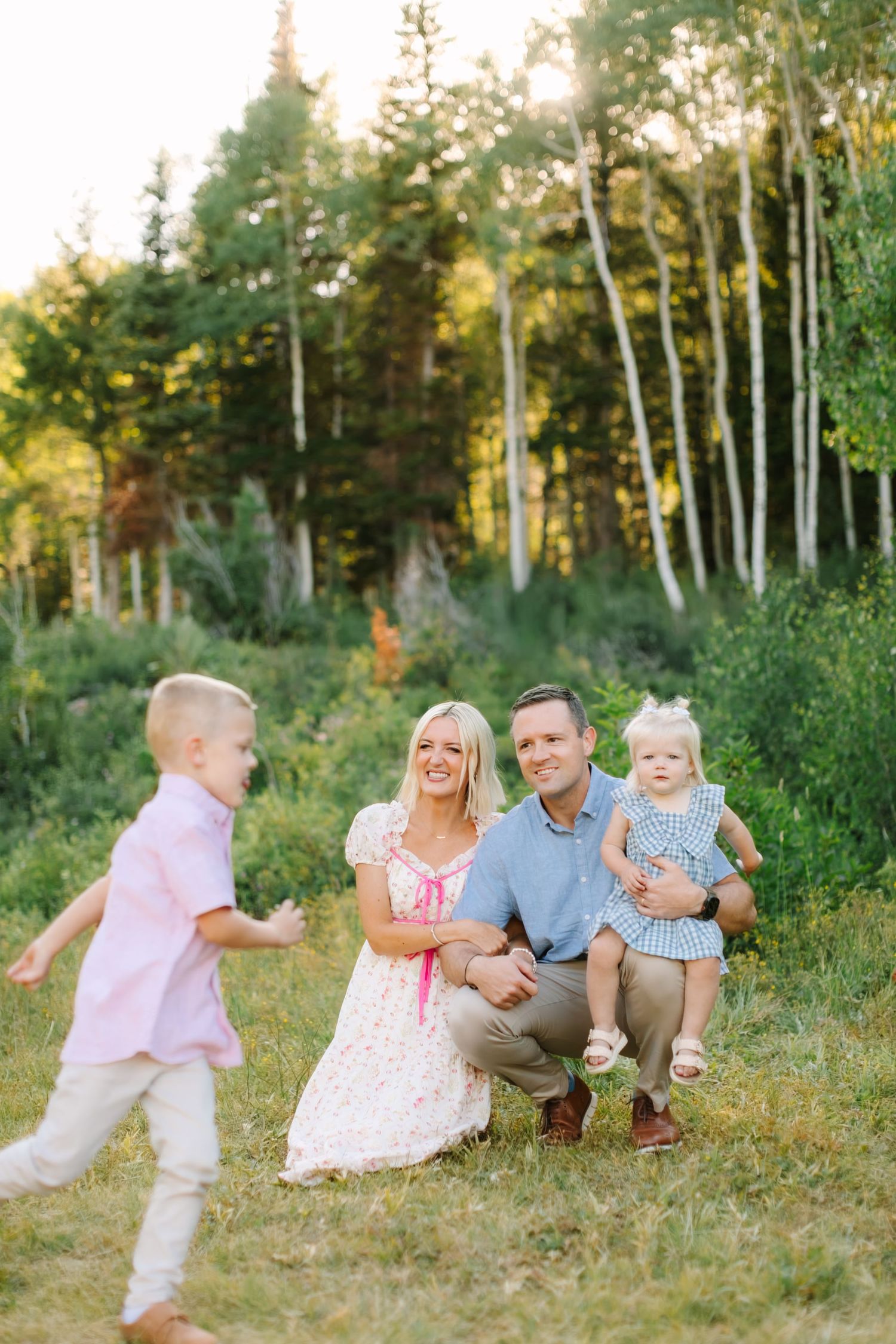 Family of four poses outdoors among aspen trees, parents crouching with toddler girl while young boy runs toward them.