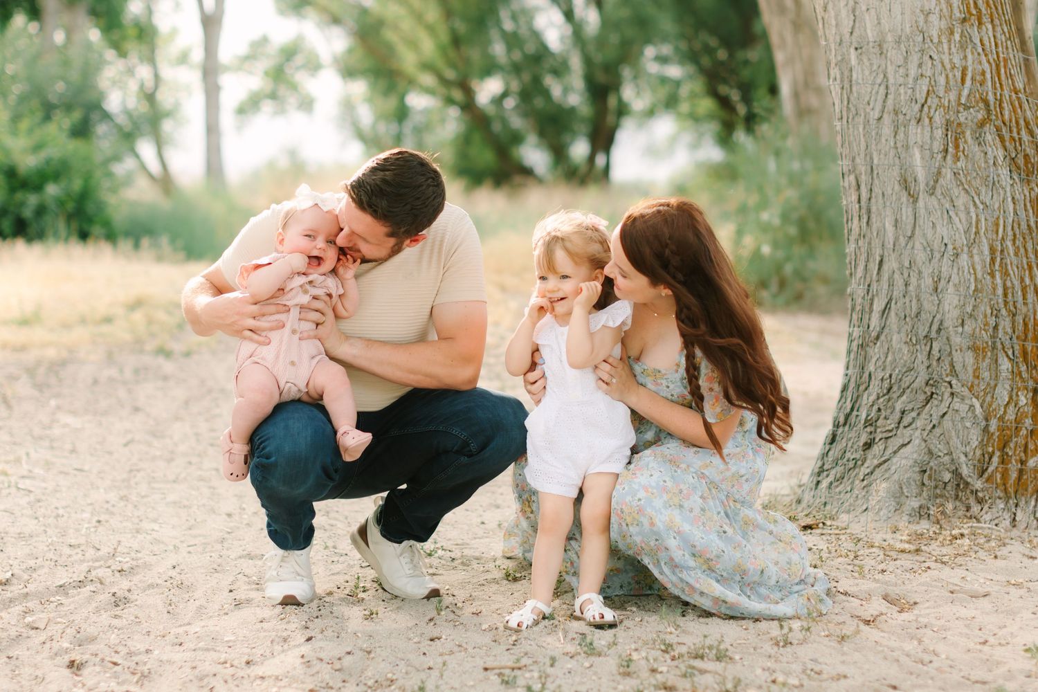 Family of four enjoying outdoor photo session near tree, parents with baby and toddler in natural setting.