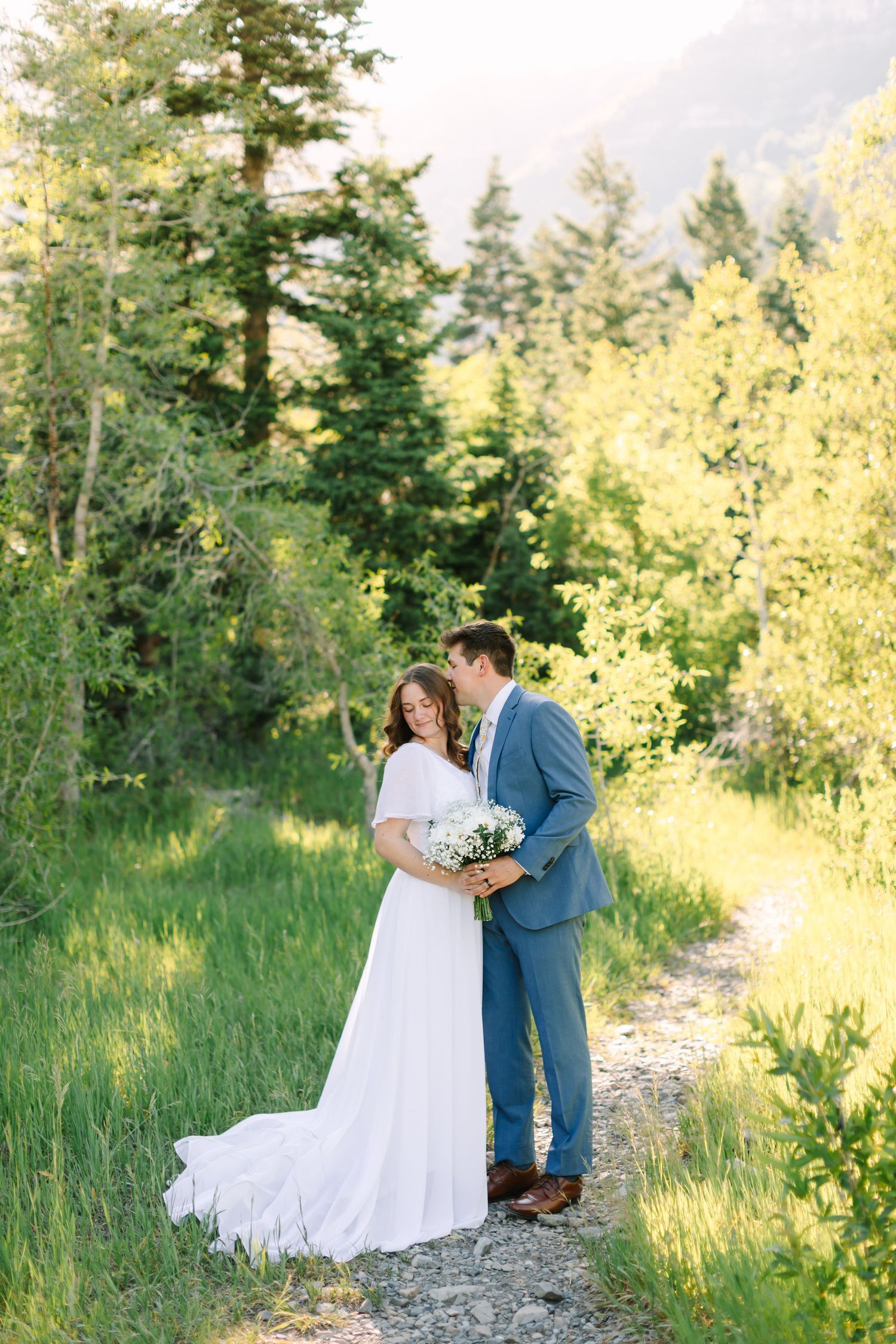 A bride and groom share a tender moment on a mountain trail surrounded by lush green trees in golden sunlight.