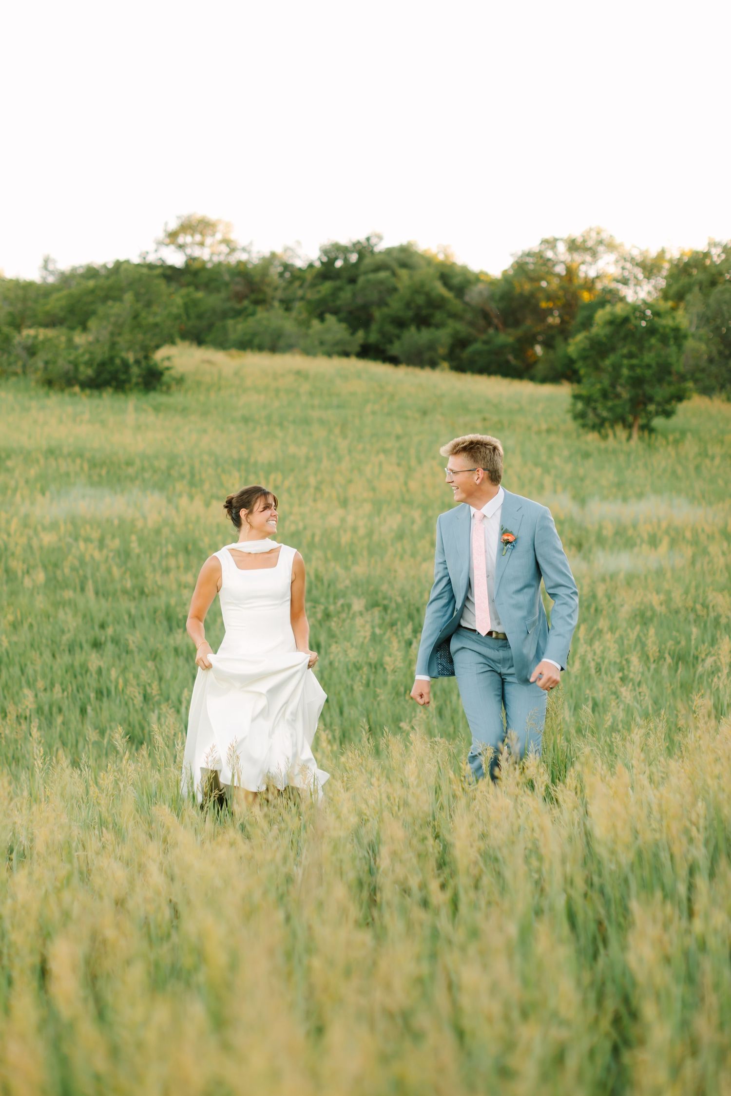 Bride and groom walking hand in hand through a lush green field during golden hour at their outdoor wedding.