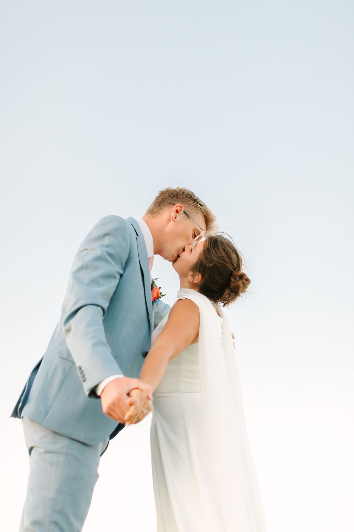 Bride and groom share a romantic kiss against a clear sky, groom in light blue suit holding bride's hand.