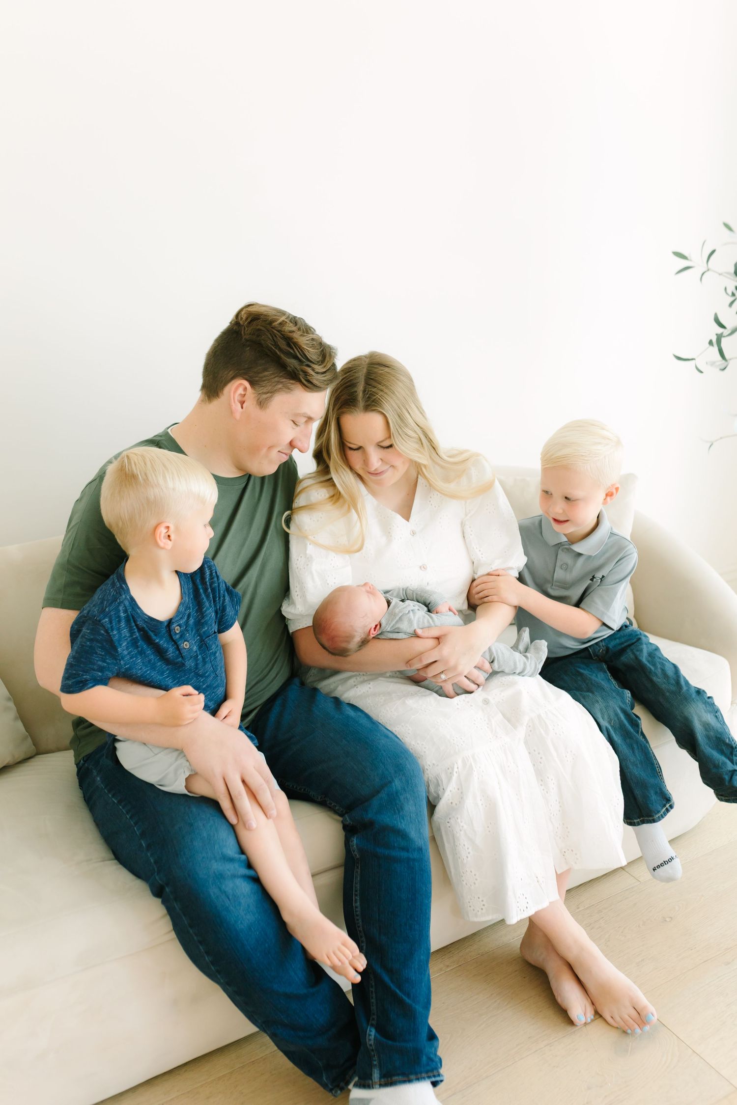 A happy family of five sits on a cream sofa, parents admiring their newborn baby while two young blonde boys smile nearby.