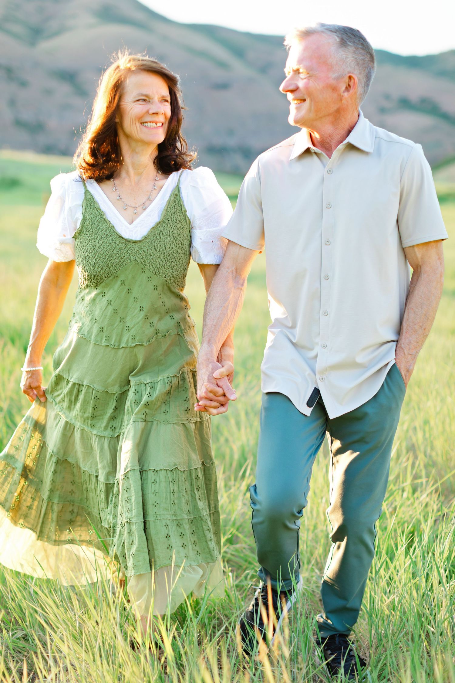 A couple walks hand in hand through a grassy meadow with mountains in the background on a sunny day.