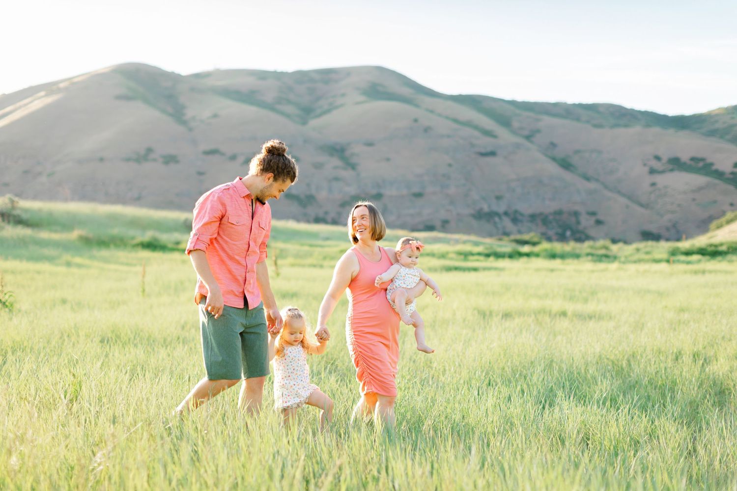 Family members walk hand in hand through a scenic green meadow with rolling hills in the background.