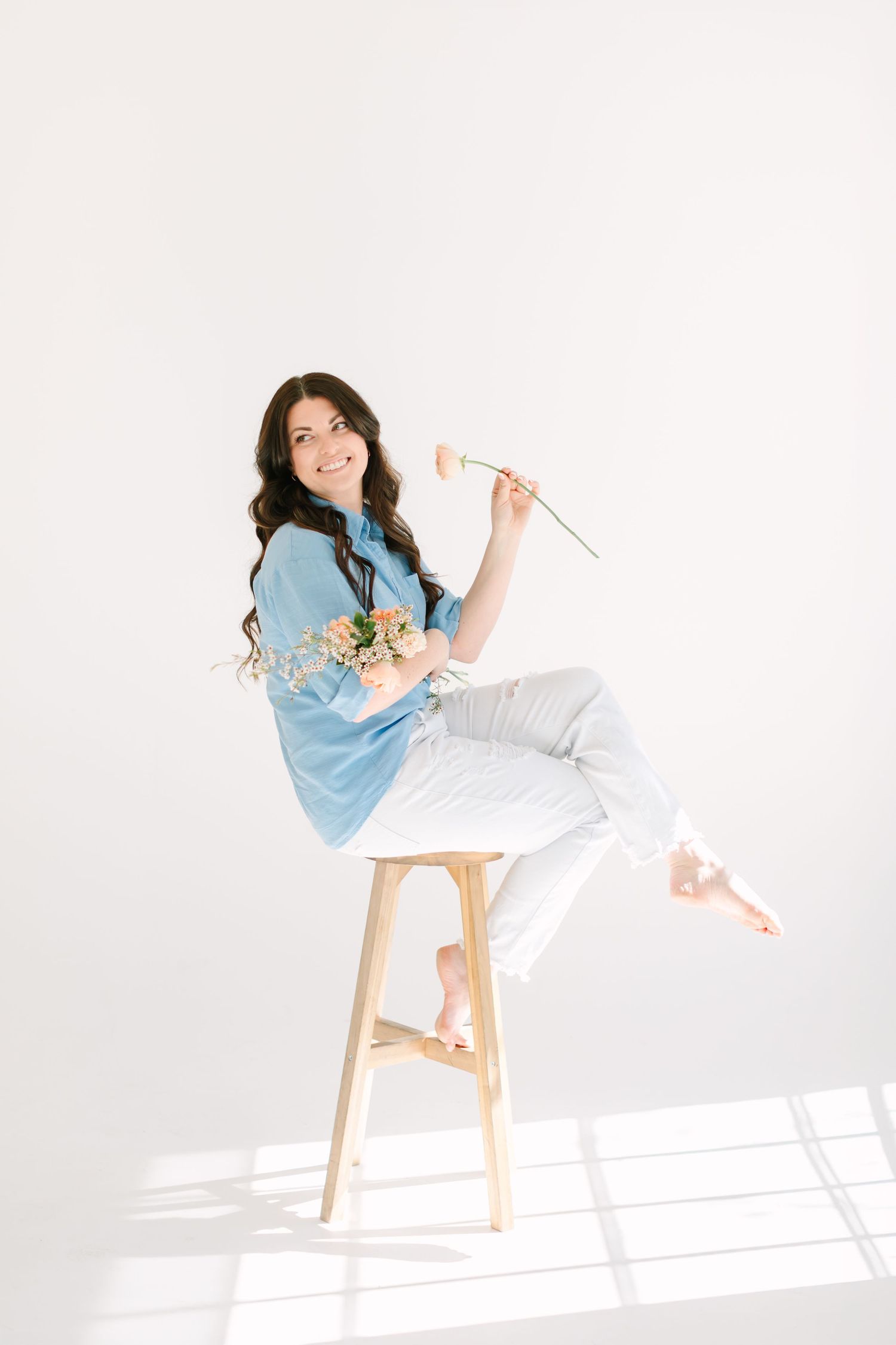 A playful pose on a wooden stool features someone in blue and white with flowers against a bright background.