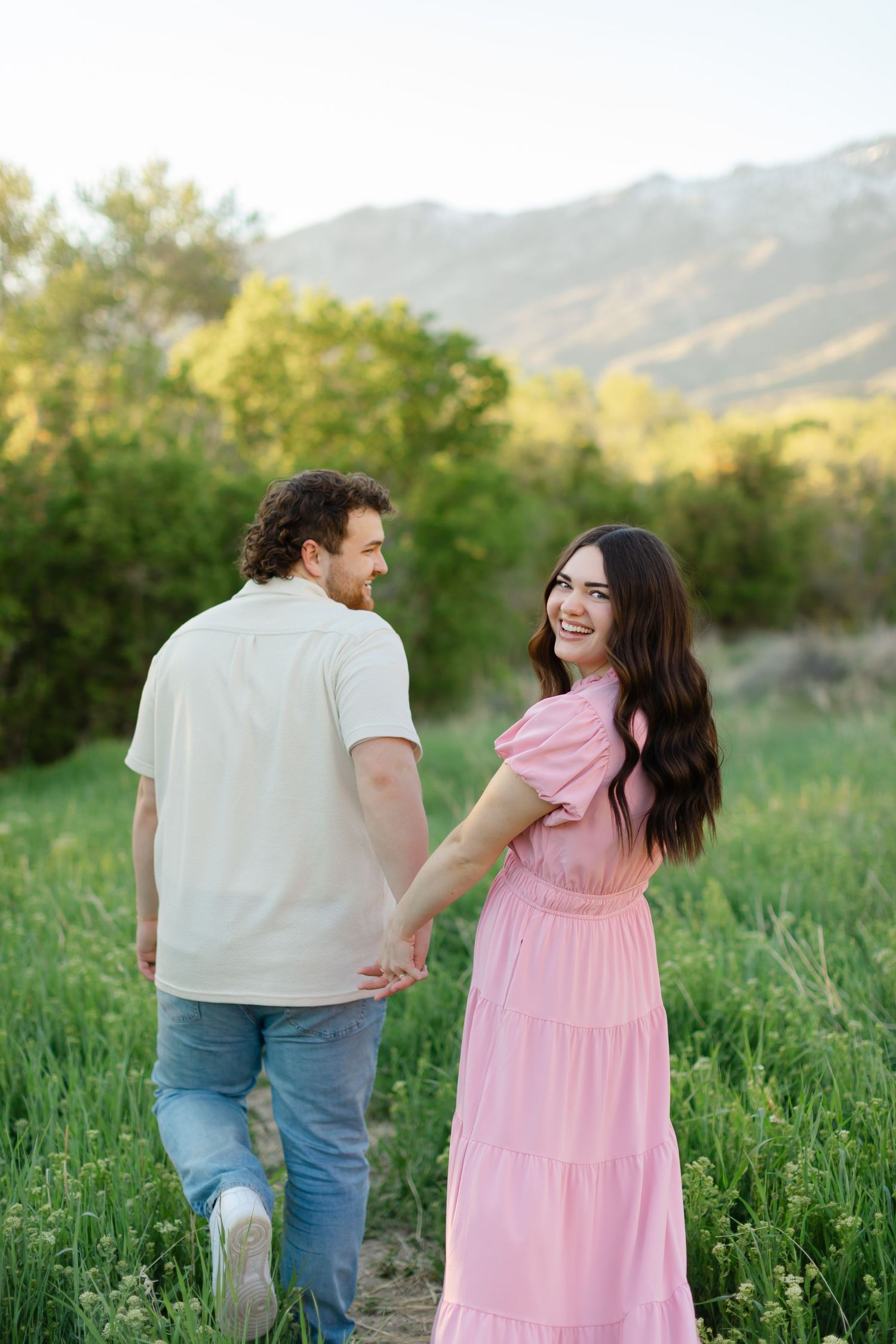 Couple walking through a green meadow at sunset with mountains in the background for their engagement photoshoot in Alpine, Utah.