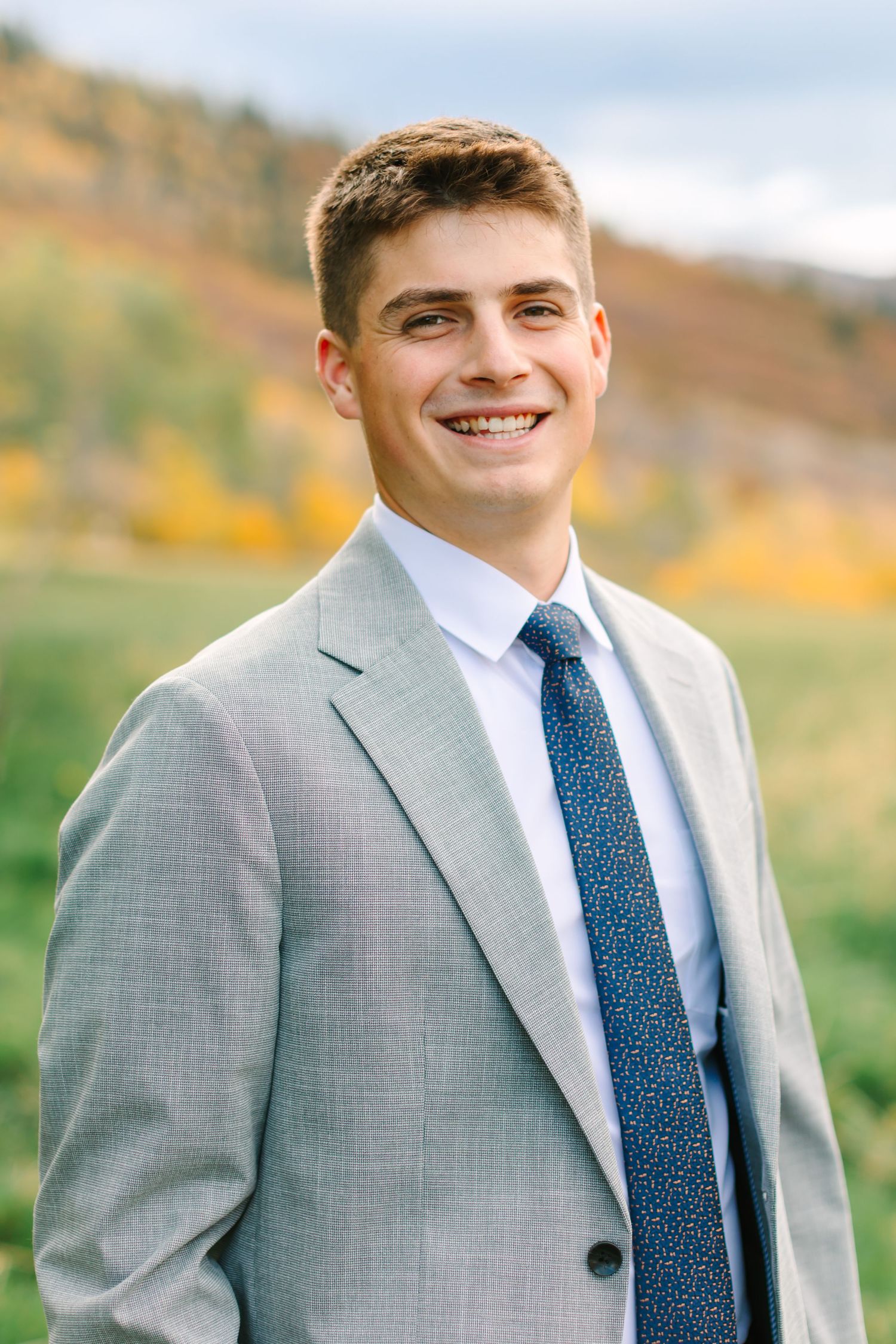 Professional portrait of someone wearing a light gray suit with navy blue tie against an autumn outdoor background.