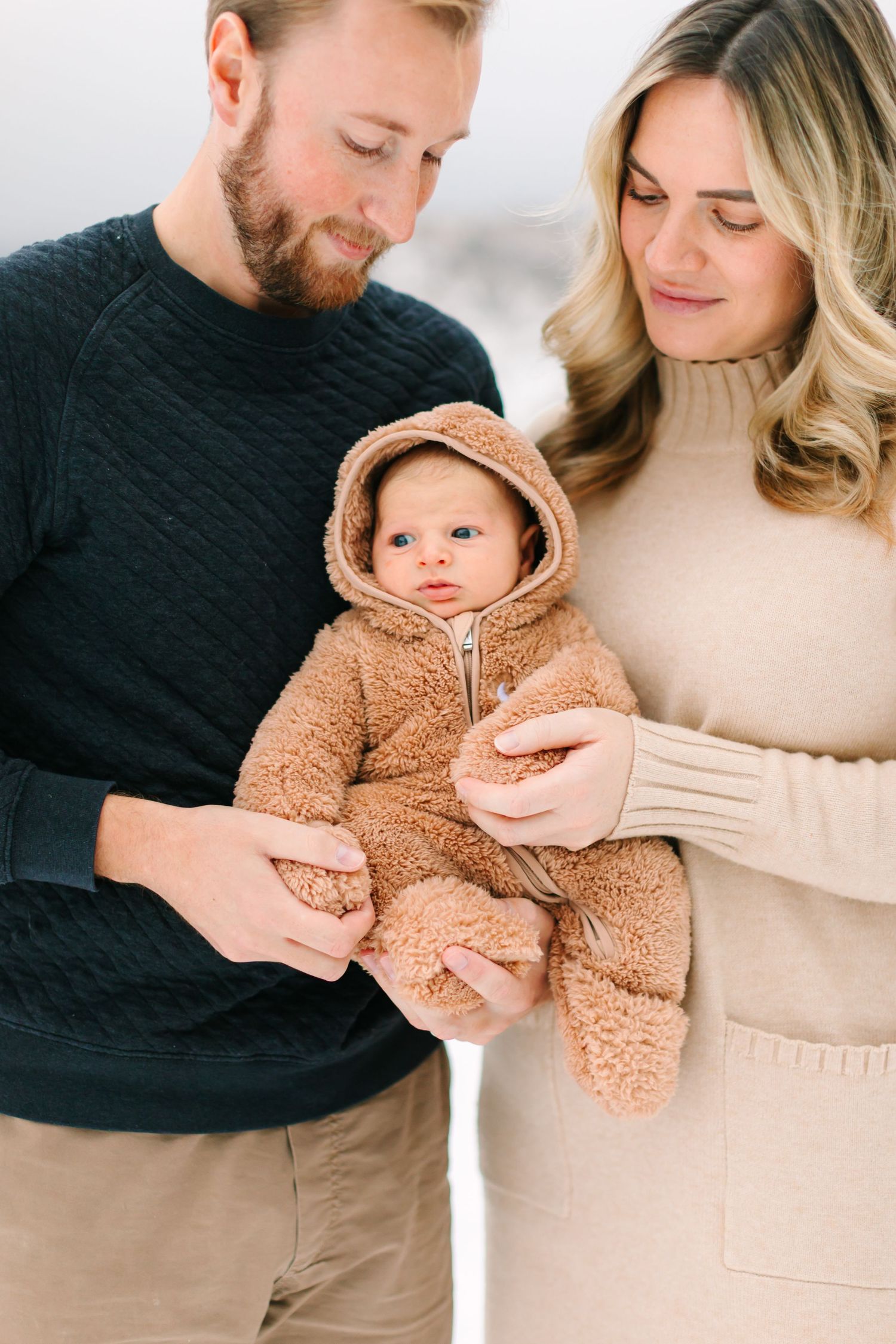 A series of tender family moments showing parents holding and interacting with their baby wearing a cozy brown teddy bear outfit.