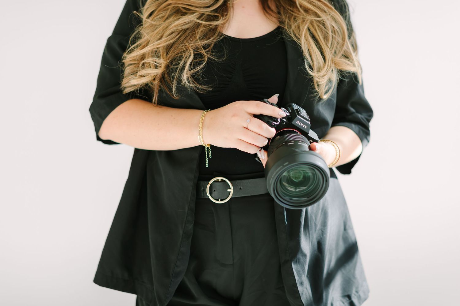 A photographer in a black outfit holds a professional DSLR camera against a white background.