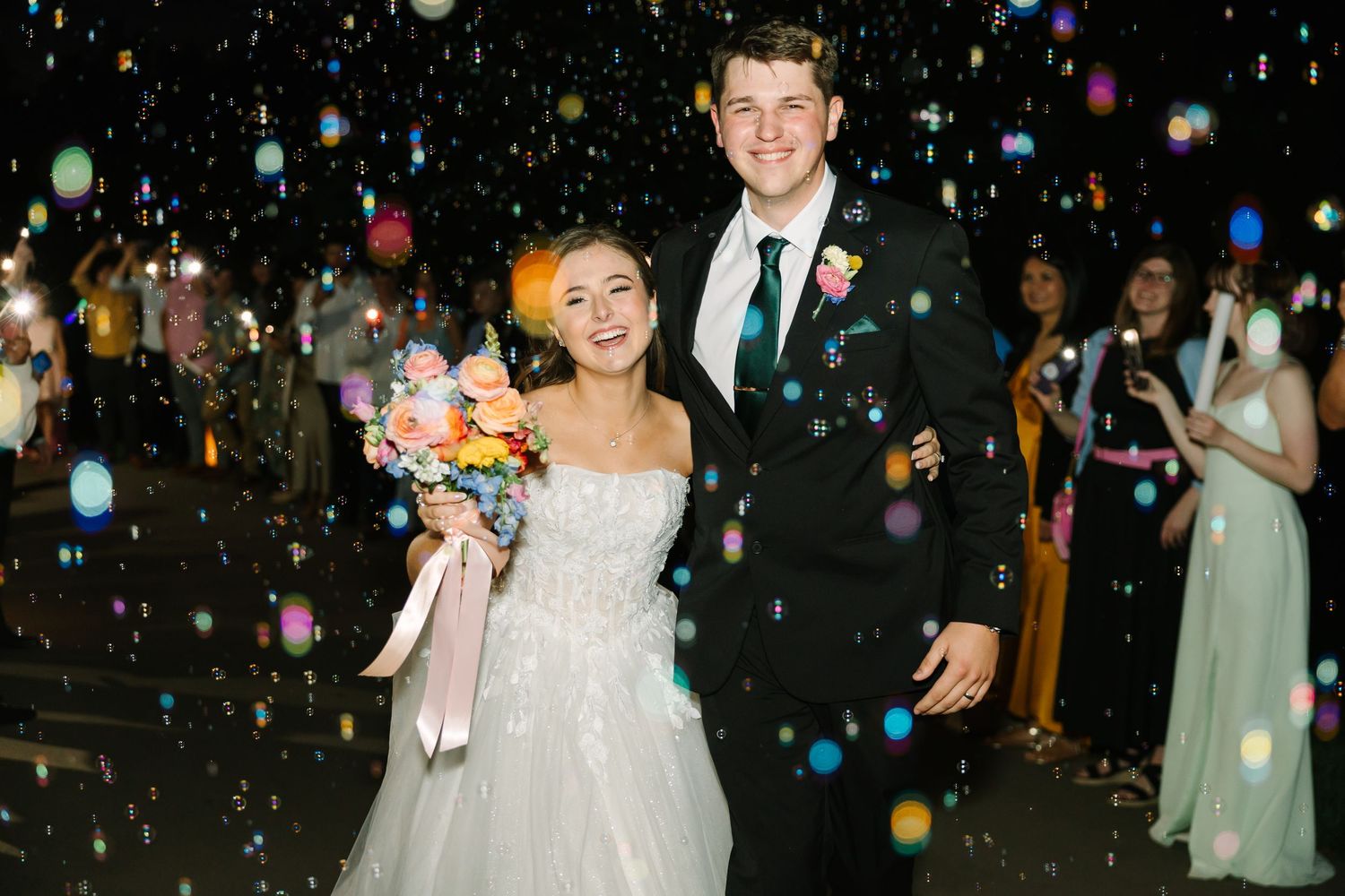 Wedding guests create a festive tunnel of glowing bubbles as newlyweds exit their reception at night.