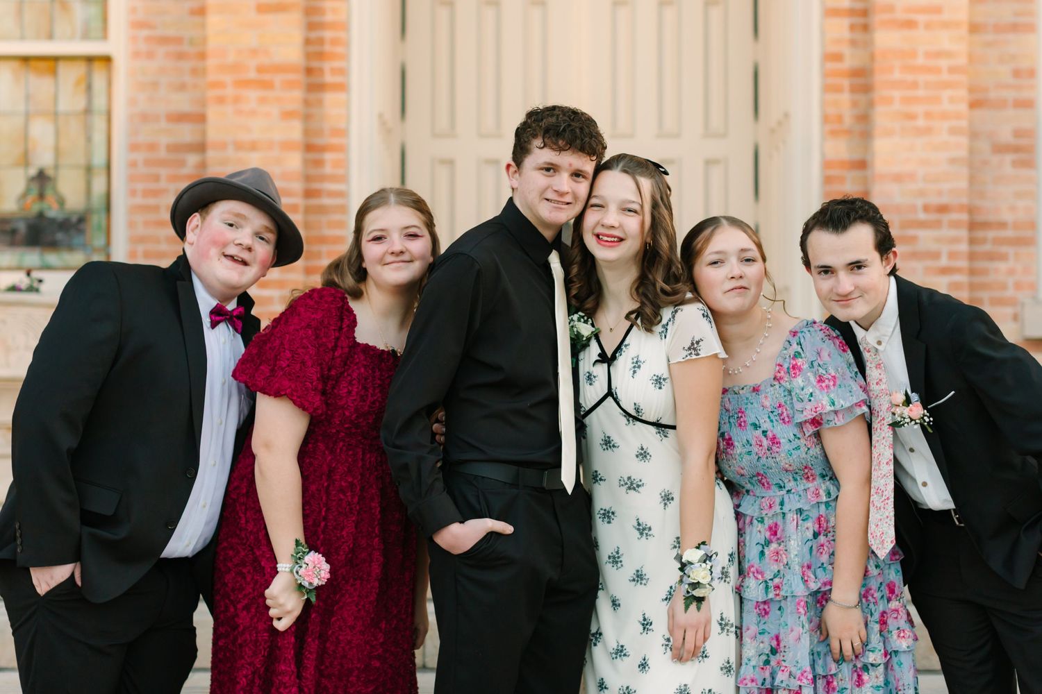 Group of people in formal attire pose together for a fun photo against a brick wall backdrop.