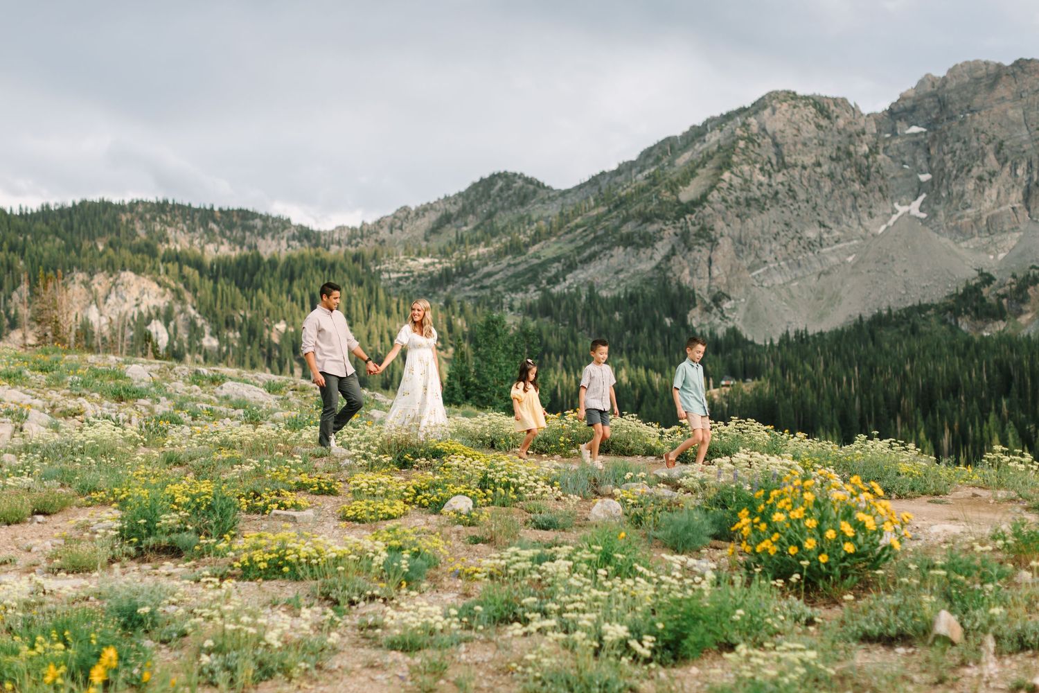 Family of four walks through wildflower meadow with mountain backdrop on a cloudy day.