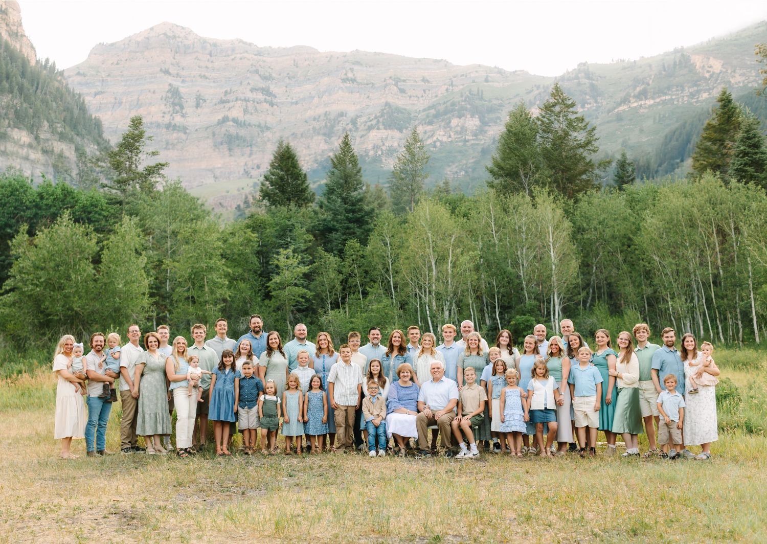 Large extended family group photo outdoors with mountain and forest backdrop on a sunny day.